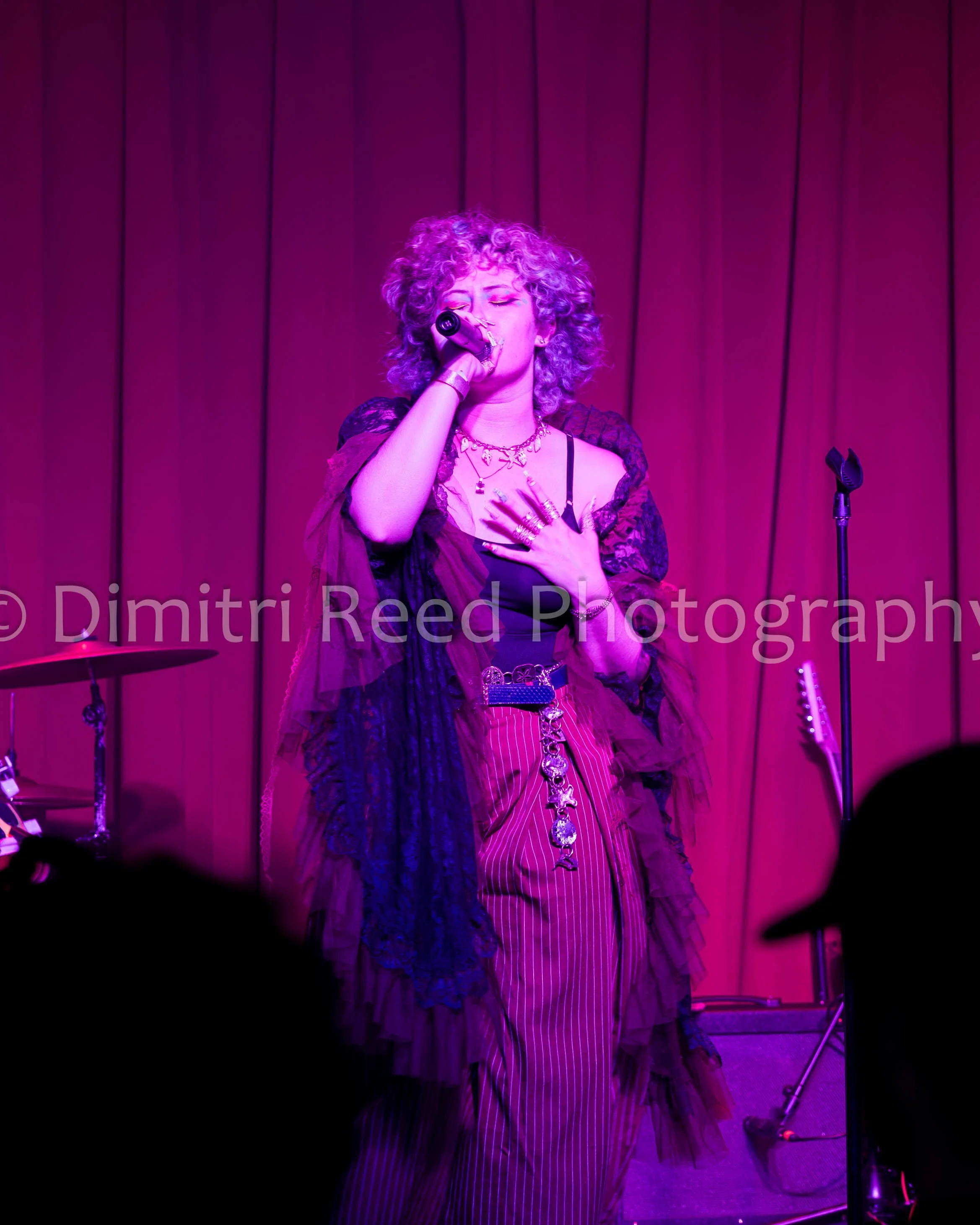 A woman with curly hair singing into a microphone on stage, illuminated by purple and pink lights, with a curtain background and drums visible.