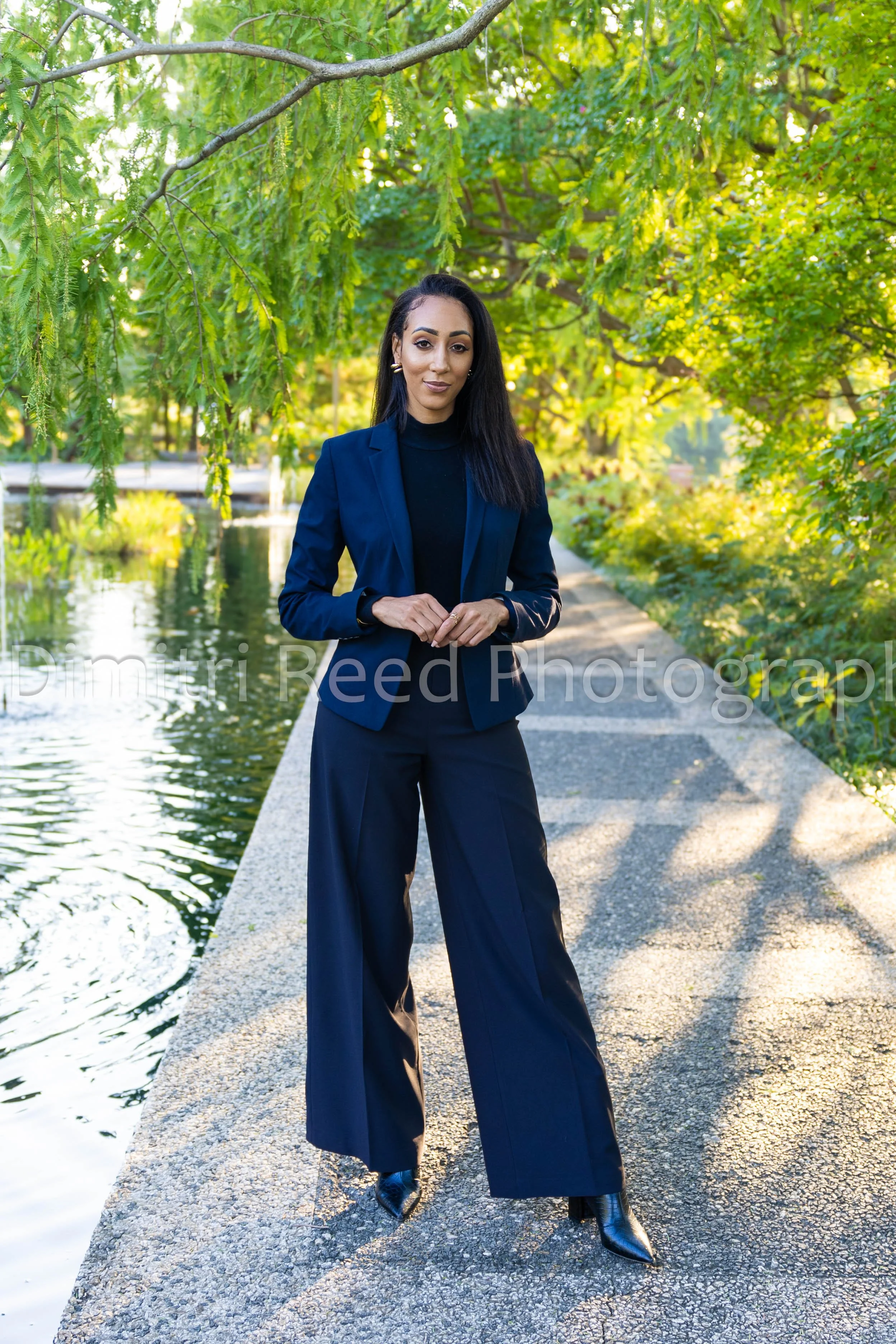 A woman in a navy blazer and wide-leg pants standing on a concrete path near a body of water, surrounded by lush green trees during daytime.