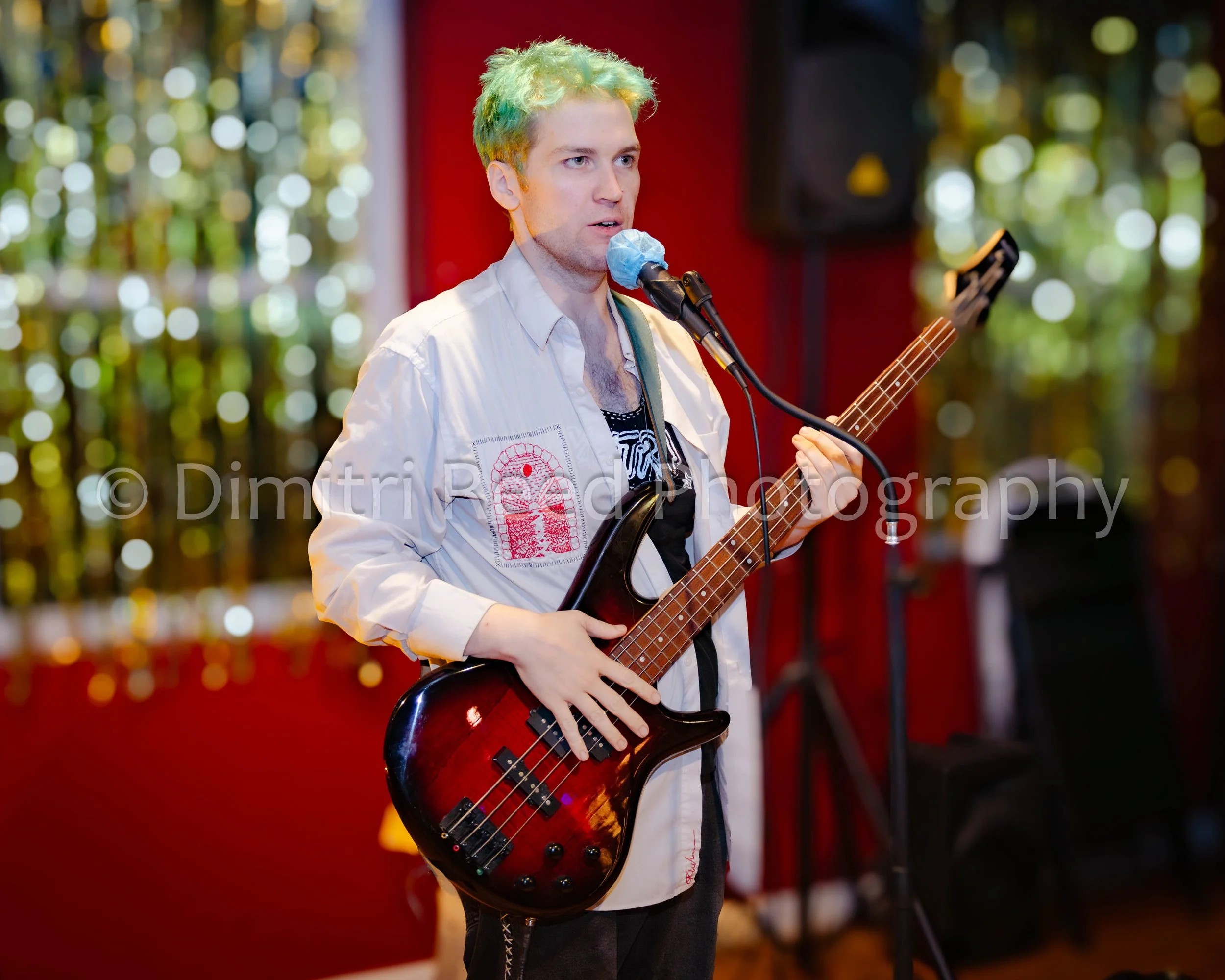 A young man with multi-colored dyed hair is playing a bass guitar and singing into a microphone. He is wearing a light-colored jacket and standing indoors with colorful, blurred background lights.