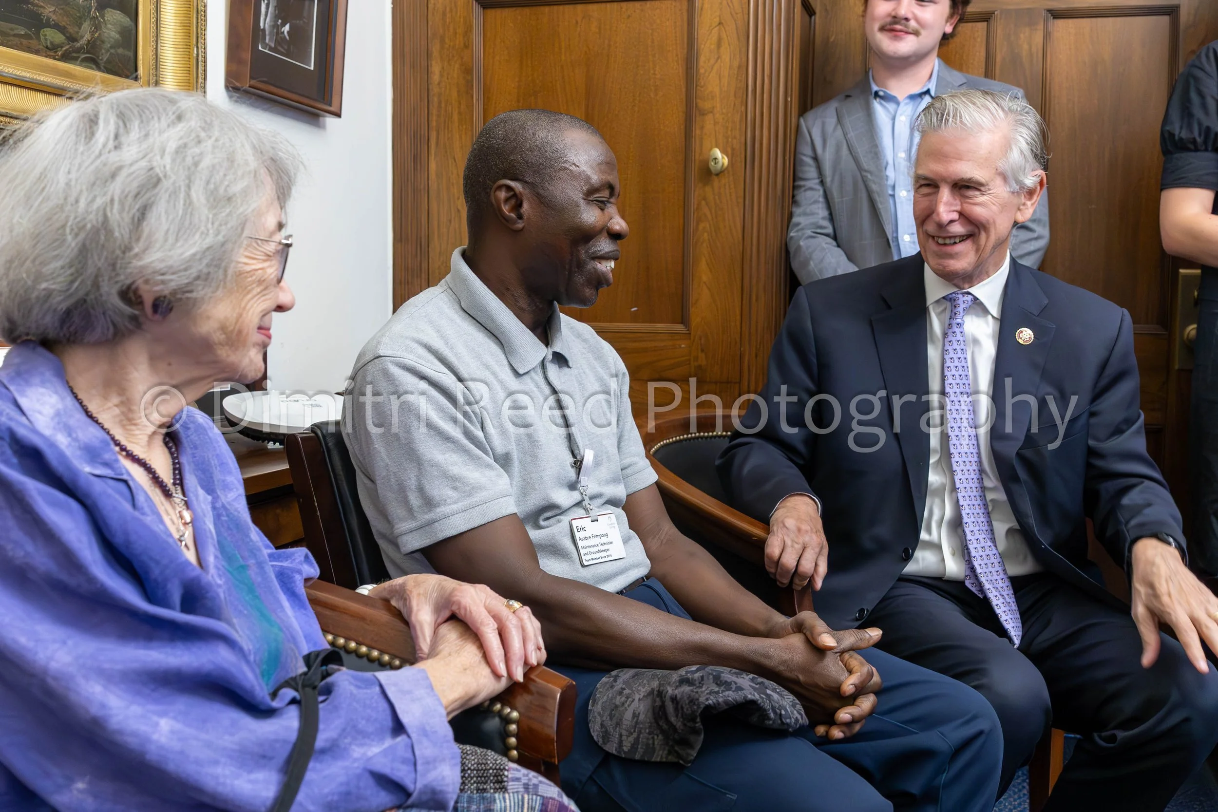Four people sitting and smiling in a room with wood-paneled walls during a discussion or meeting. One woman with gray hair and glasses, one man with a shaved head and a lanyard, another man with gray hair and a suit, and a young man standing in the b