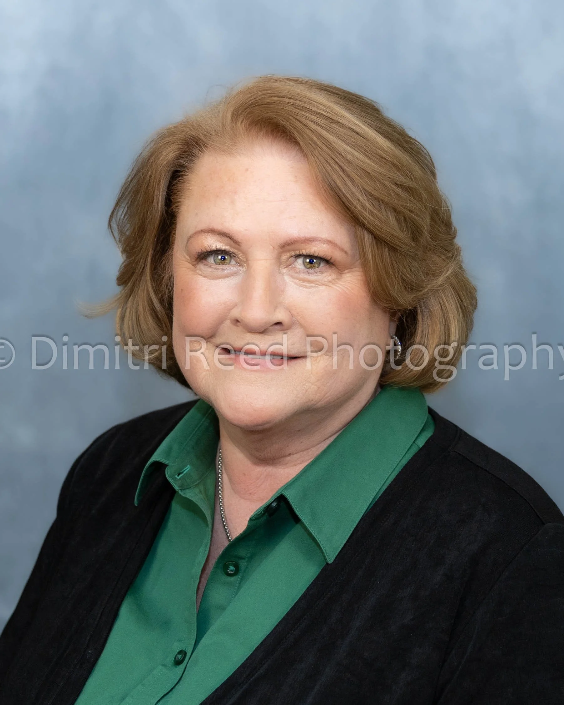 Close-up of a smiling middle-aged woman with red hair and green eyes, wearing a green collared shirt and black cardigan, standing against a plain blue background.