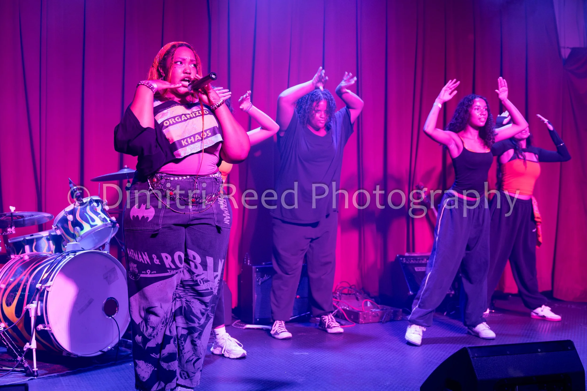 A group of women singing or performing on a stage with red curtains, purple lighting, and musical instruments nearby.