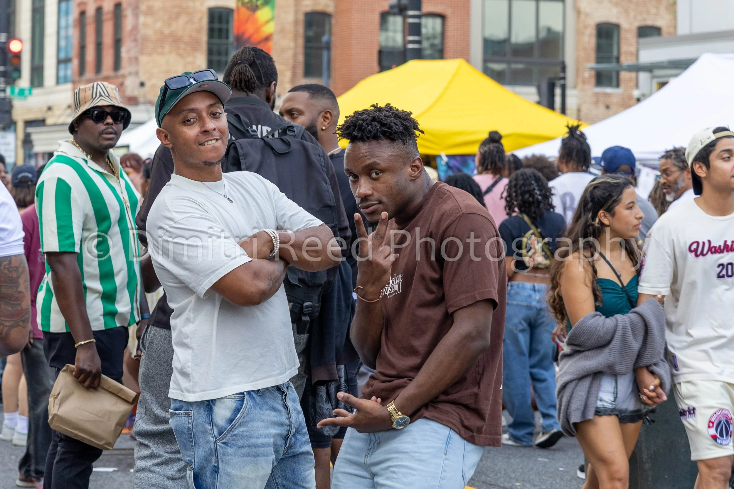 Two men are posing in a crowded outdoor event, with one giving a fist bump to the other. The man on the left is wearing a white t-shirt, jeans, and a green cap with sunglasses on top. The man on the right is wearing a brown t-shirt, jeans, and a gold
