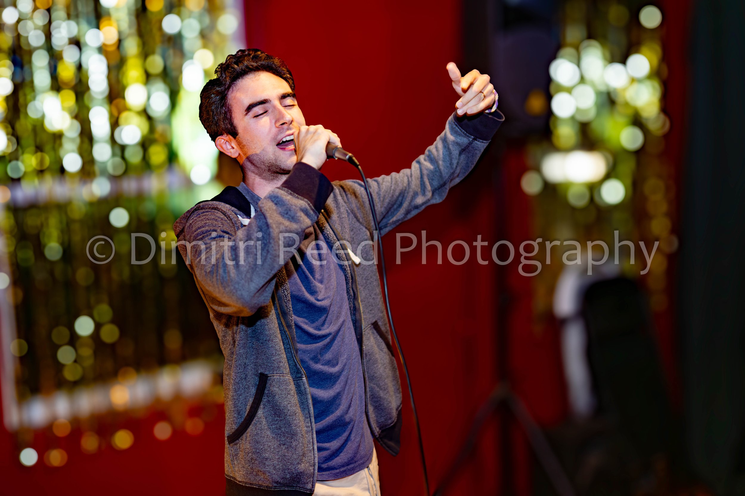 Young man singing passionately into a microphone with eyes closed, holding the microphone with one hand and gesturing with the other, in front of a blurred background of colorful lights.