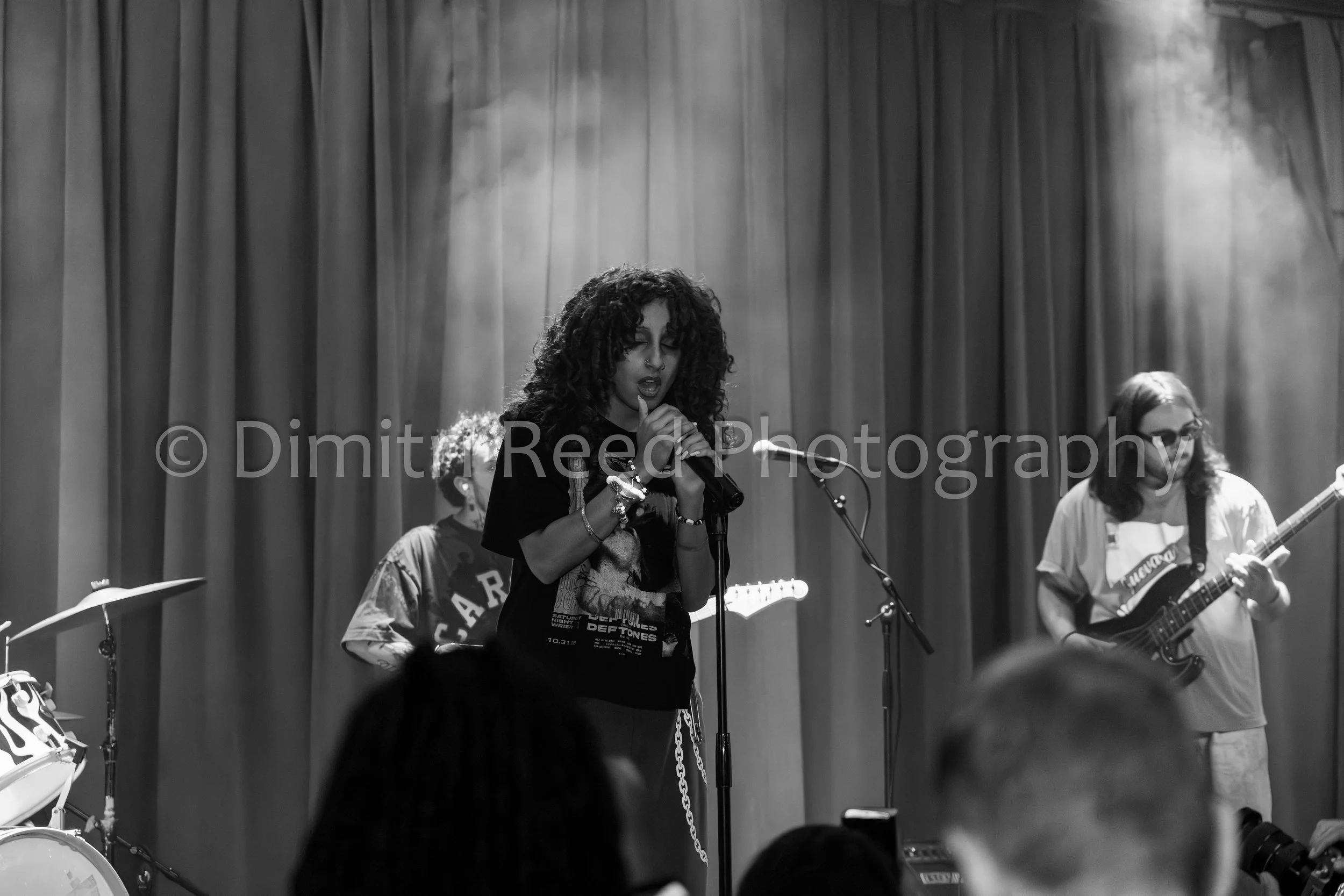 A black-and-white photo of a female singer with curly hair performing with a microphone on stage, surrounded by band members playing instruments, in front of a curtain.
