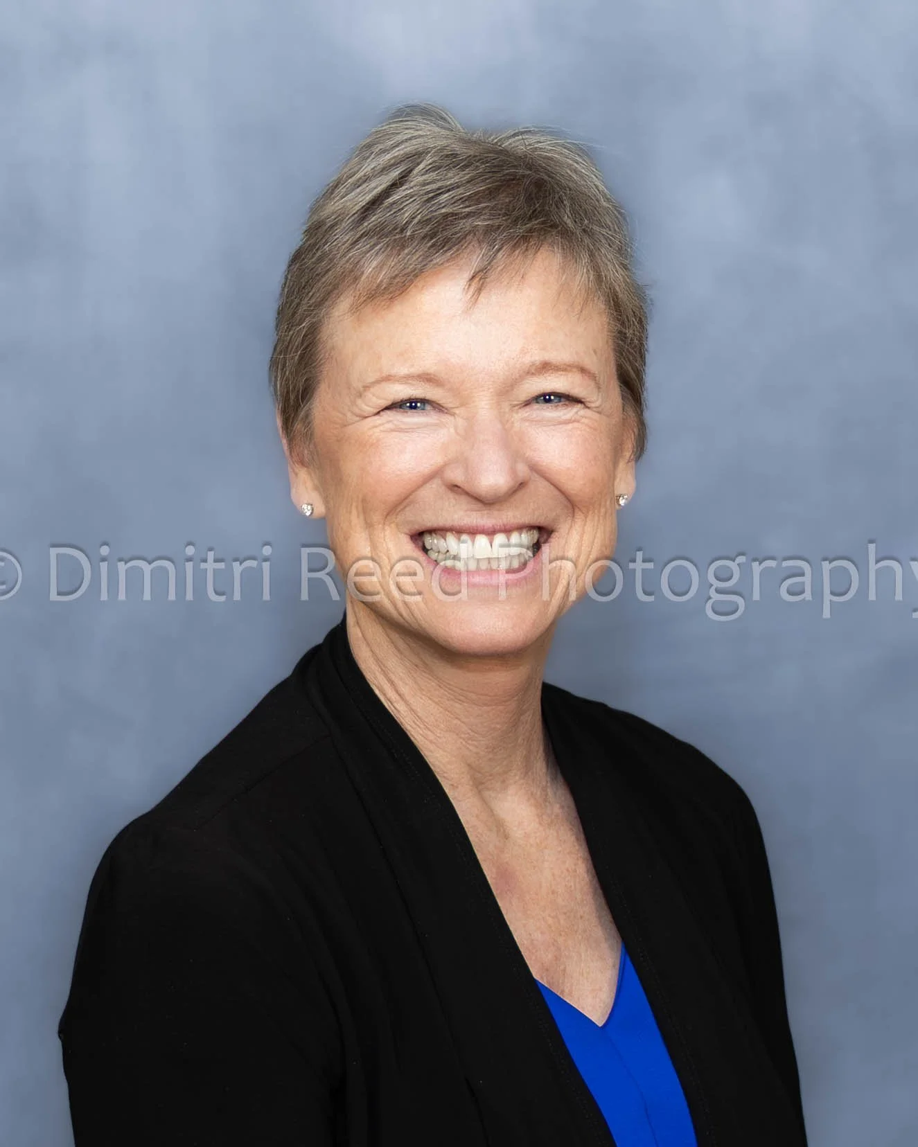 A smiling middle-aged woman with short hair wearing a black blazer and blue top against a gray background.