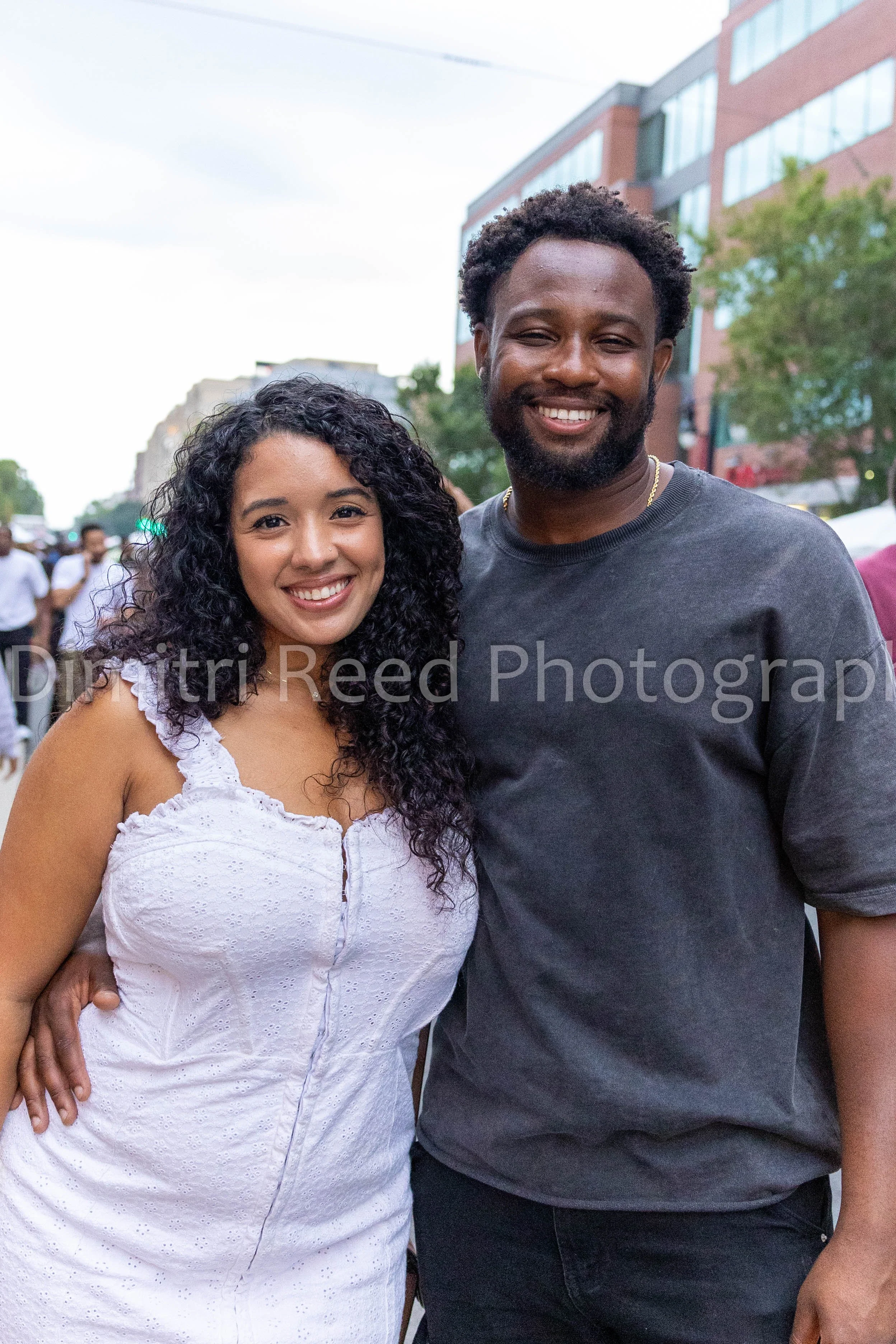 Smiling woman with curly hair wearing a white dress and smiling man with a beard wearing a black t-shirt standing outdoors with a city street, trees, and buildings in the background.