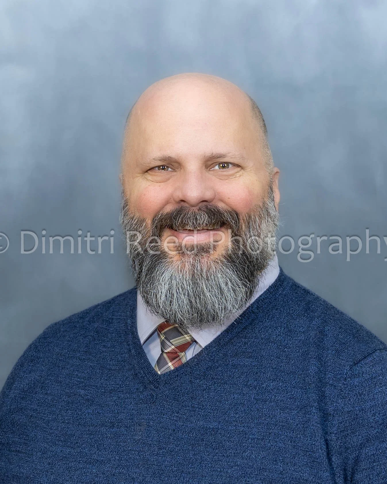 A middle-aged man with a bald head, a full beard, and a smile, wearing a blue sweater, a checked tie, and a light-colored shirt. Background is a plain grayish-blue color.