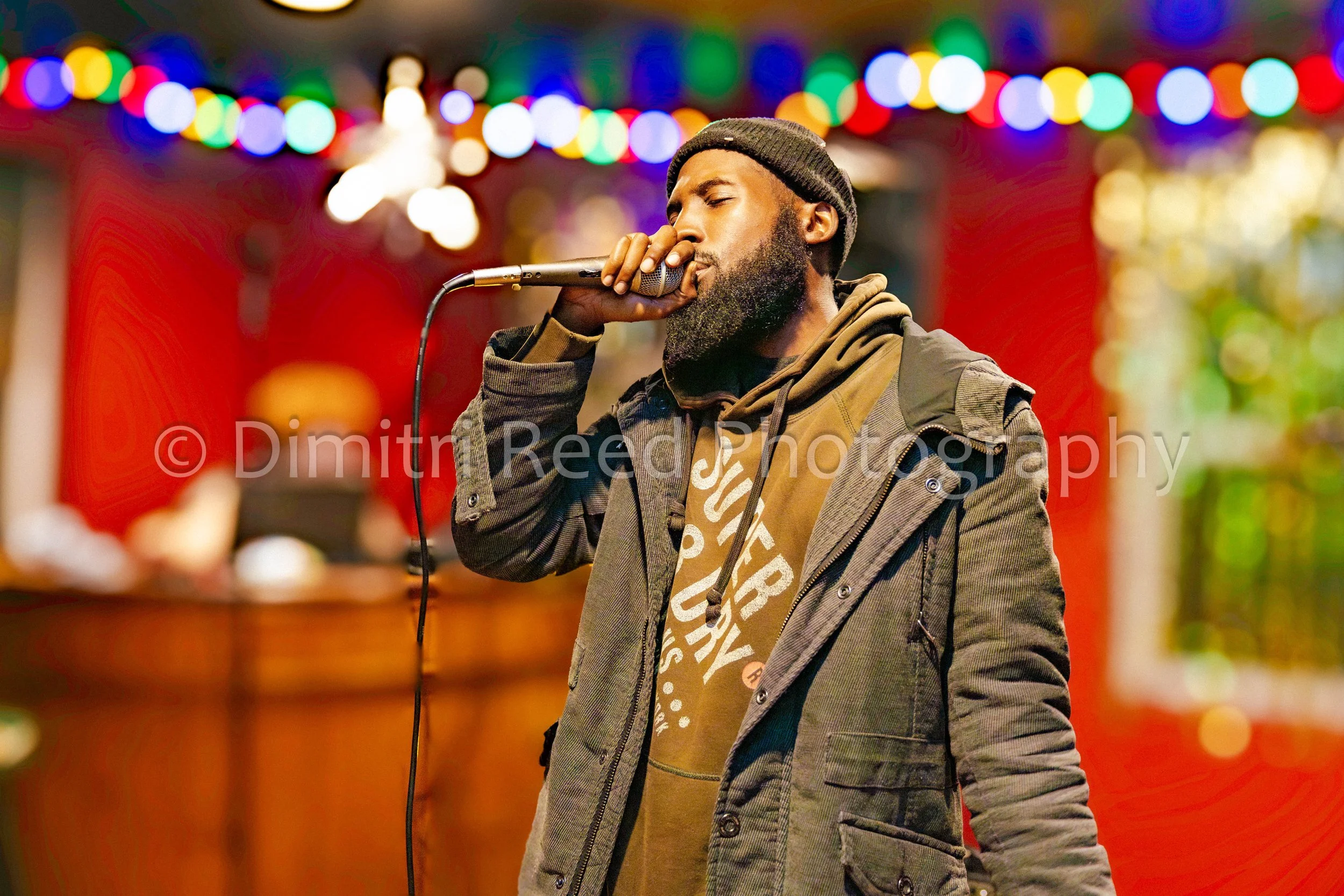 A man singing into a microphone with colorful blurred lights in the background, wearing a black beanie, hoodie, and jacket.