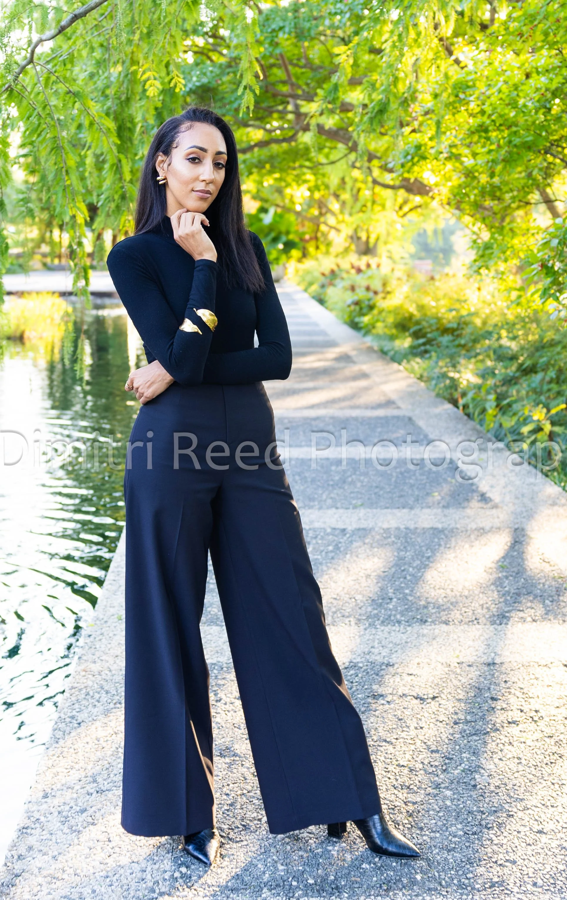 A woman wearing a black turtleneck, black wide-leg pants, and black high-heeled boots stands on a gravel pathway near a lake, surrounded by green trees with sunlight filtering through.