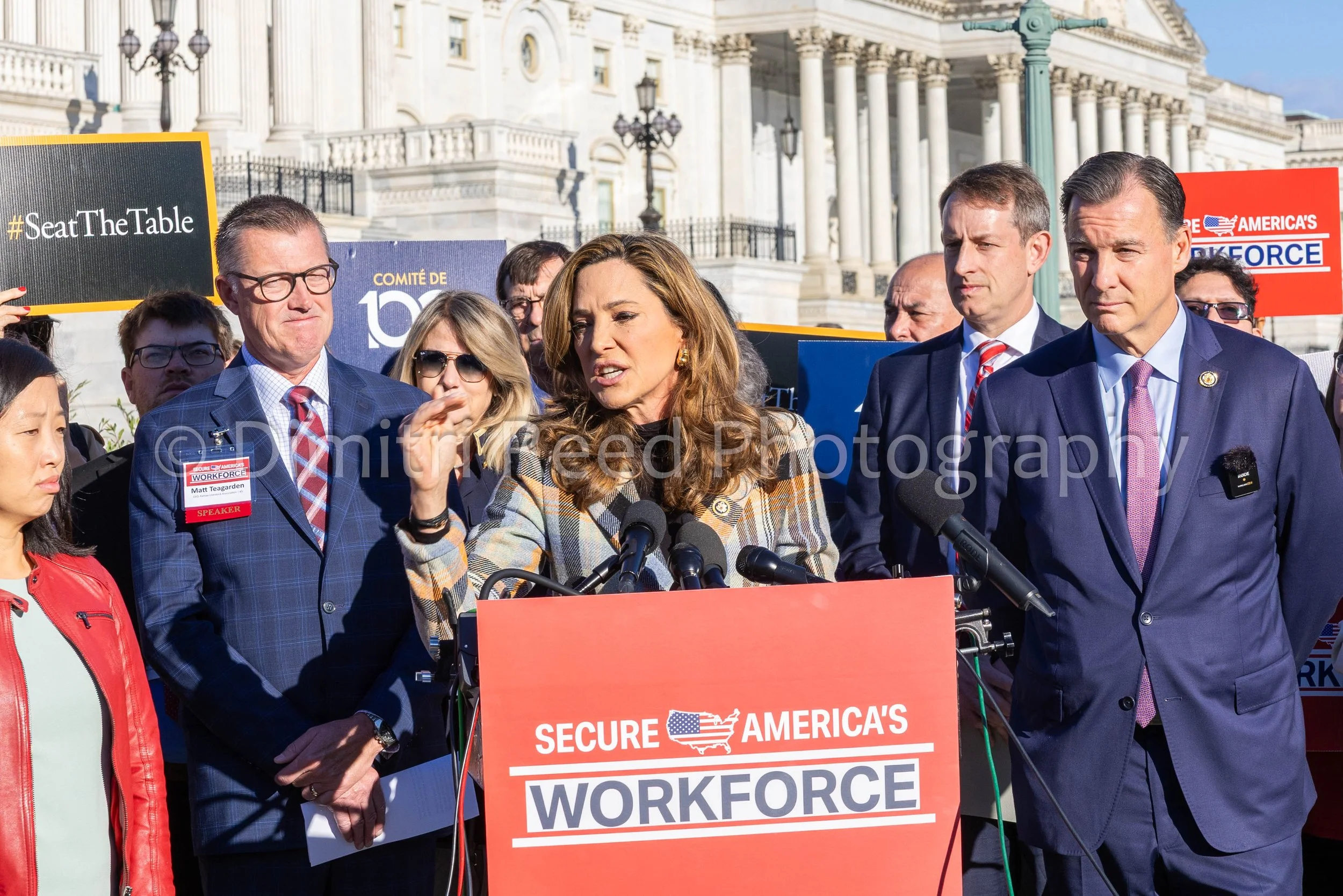 A group of people, including a woman speaking at a podium, standing outside the Capitol building. They are holding signs and wearing business attire, supporting a political rally.