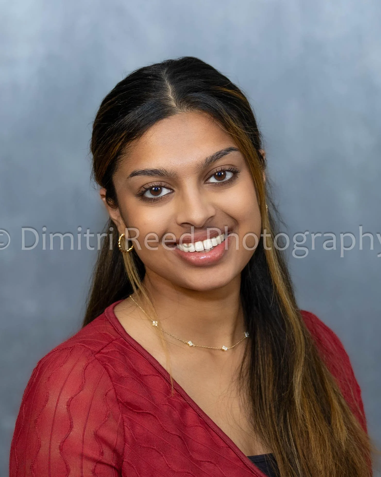 A young woman with long brown hair and brown eyes, smiling, wearing a red top and gold jewelry, against a neutral background.