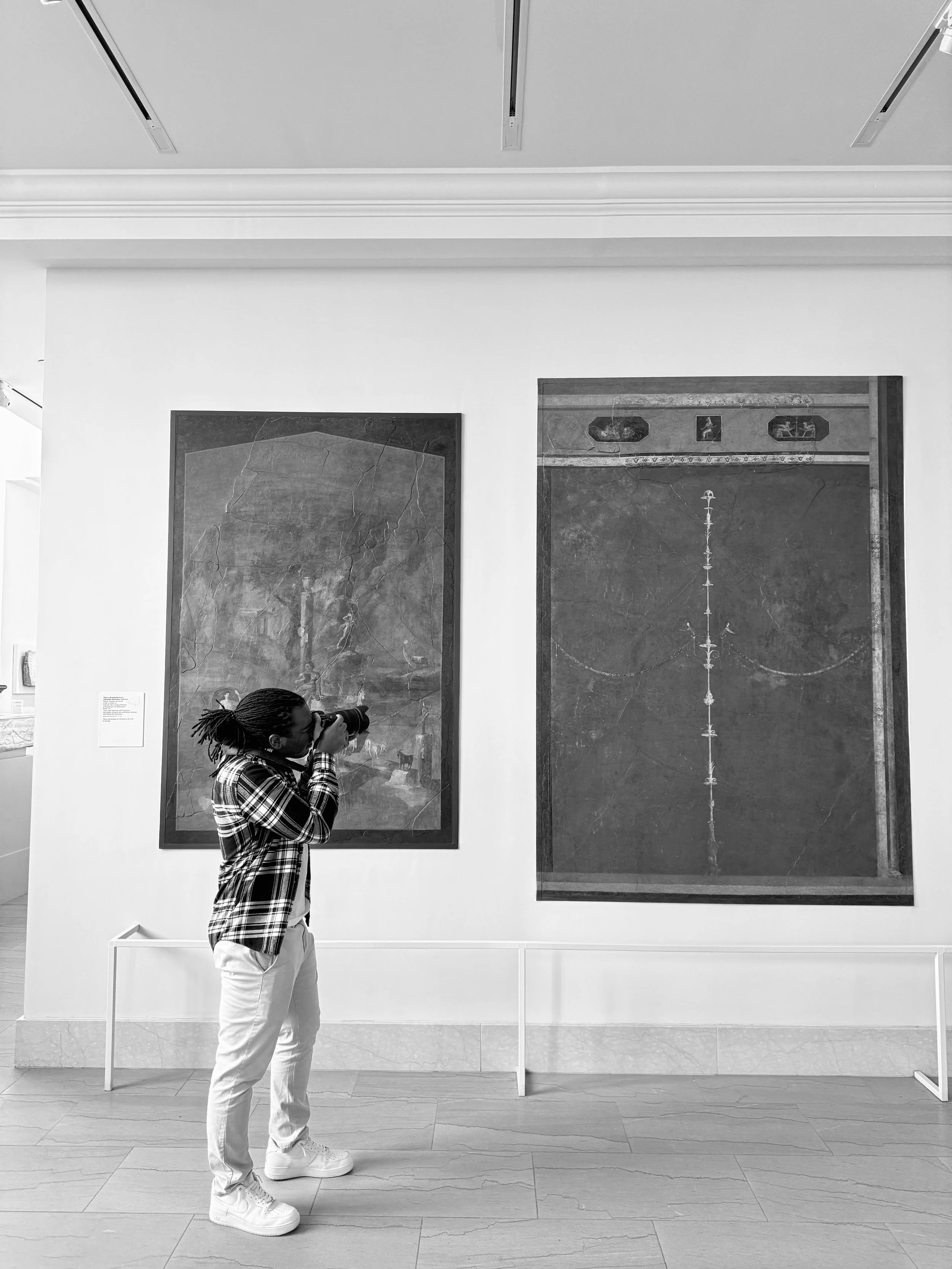 A person with braided hair wearing a plaid shirt and white pants taking a photo with a camera at an art gallery, standing in front of two large framed paintings on a white wall.