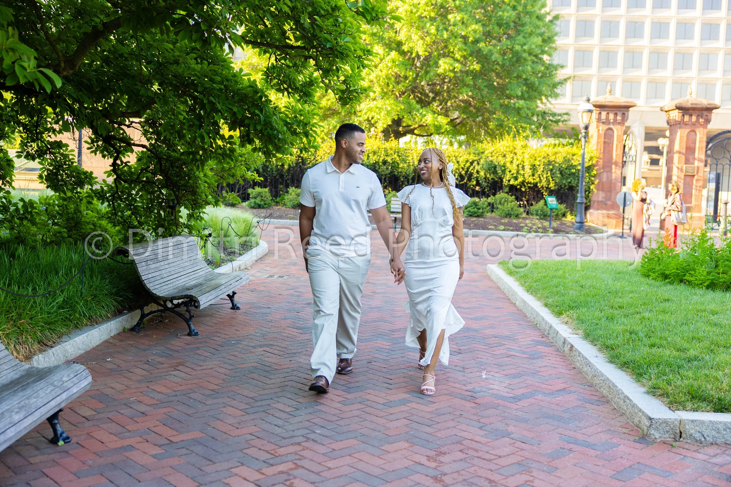 A man and woman walking hand in hand on a brick path in a park, smiling and looking at each other. The man is wearing a white polo shirt and light-colored pants, and the woman is wearing a white dress with ruffled sleeves and high heels. Green trees 
