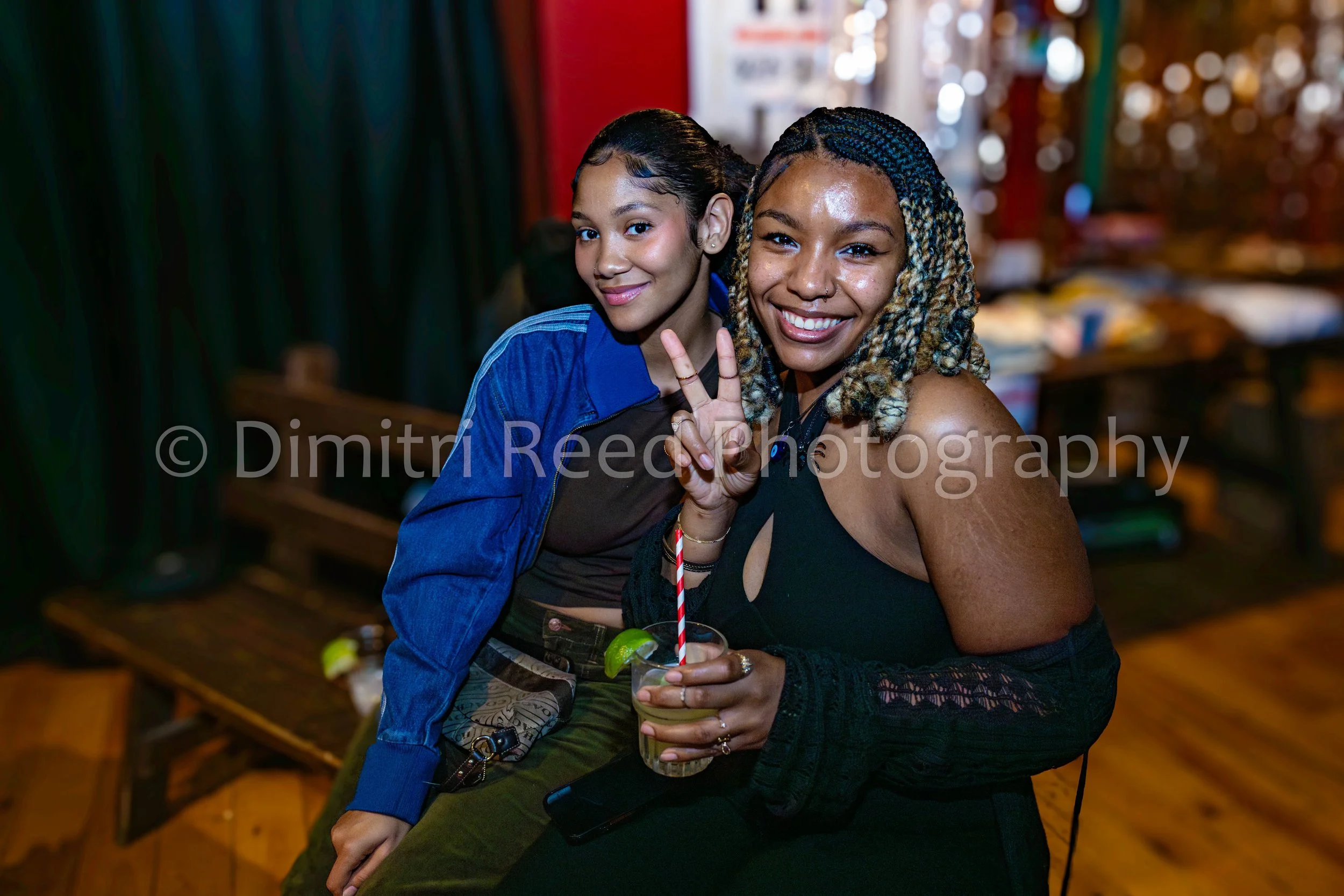 Two women sitting together in a bar or club, smiling at the camera. One woman holds a drink decorated with lime slices and a striped straw. The woman on the right makes a peace sign with her fingers. The background has colorful blurred lights and she