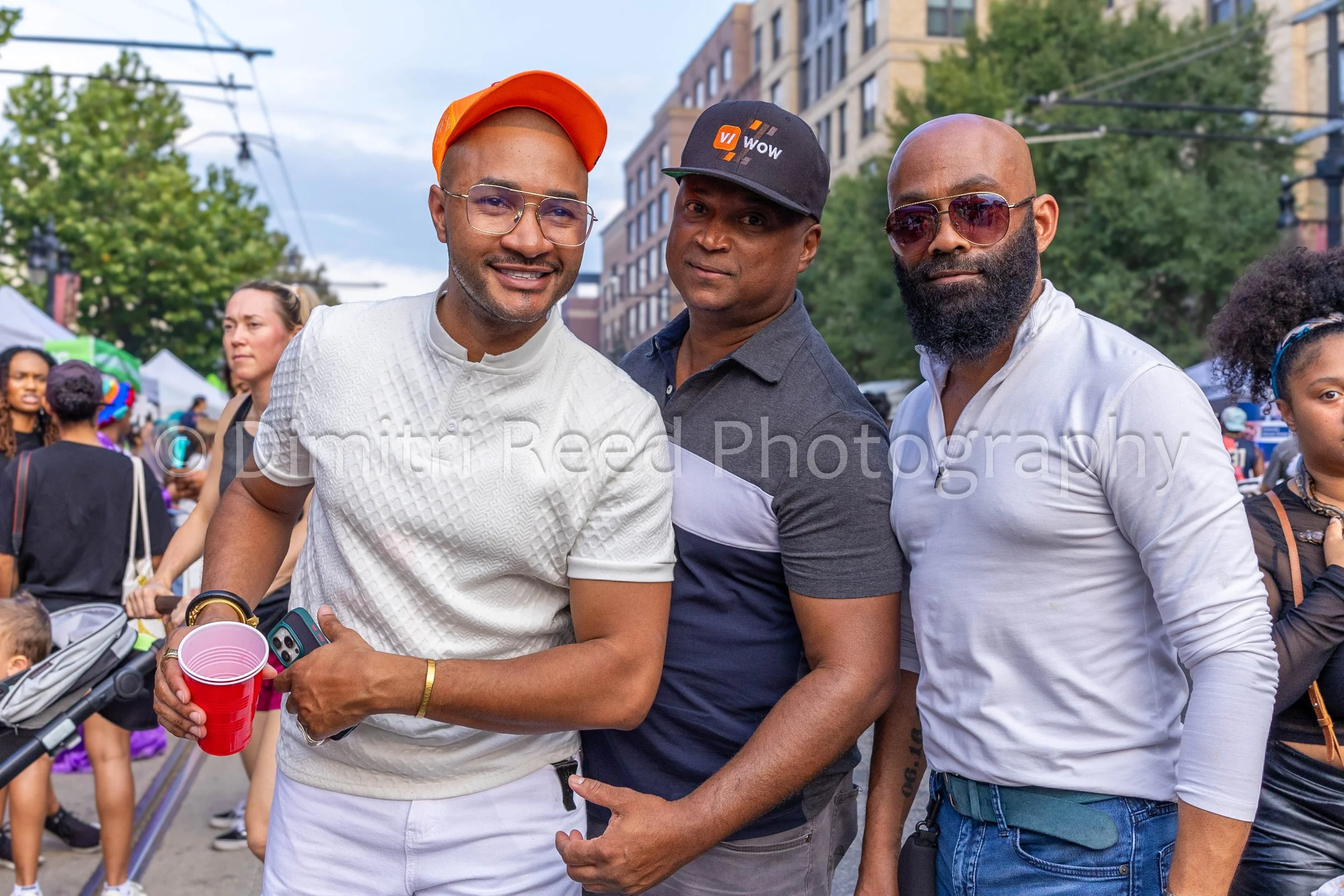 Three men standing together at an outdoor event, with people and tents in the background. The man on the left is smiling and holding a red cup, wearing glasses, a white textured shirt, and an orange cap. The man in the middle is wearing a black cap w
