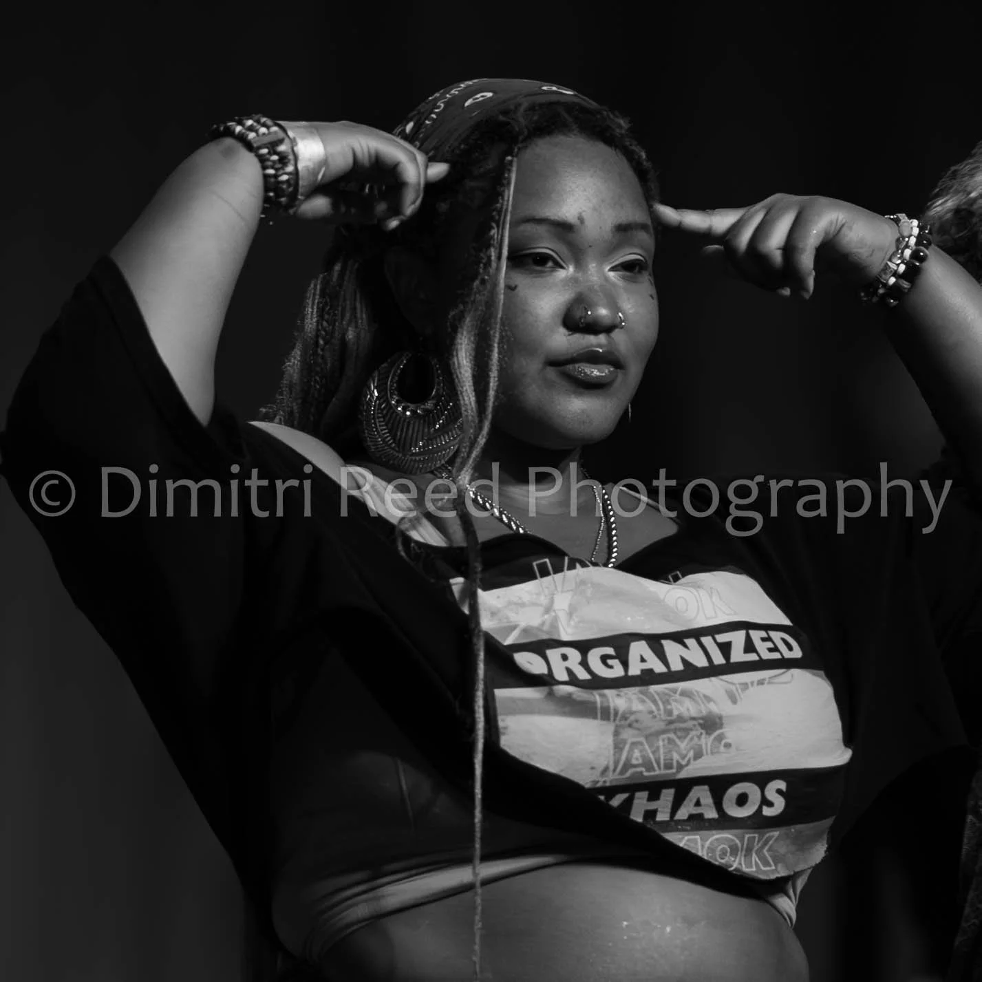 Black and white photo of a woman with various jewelry and accessories, holding her hair back, looking at the camera with a confident expression, wearing a shirt with text that includes 'ORGANIZED' and 'CHAOS'.