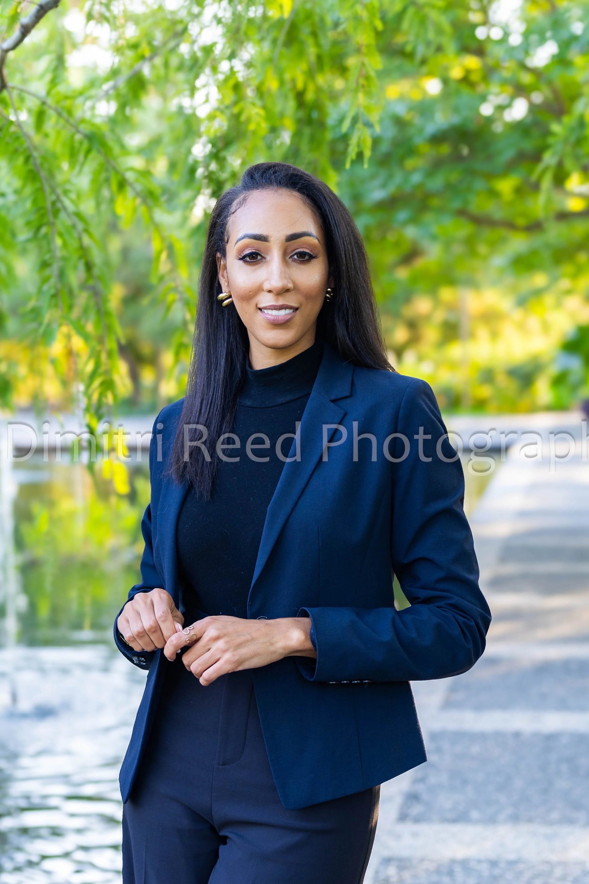 A woman in a dark blazer and pants standing outdoors near a body of water with green trees in the background.