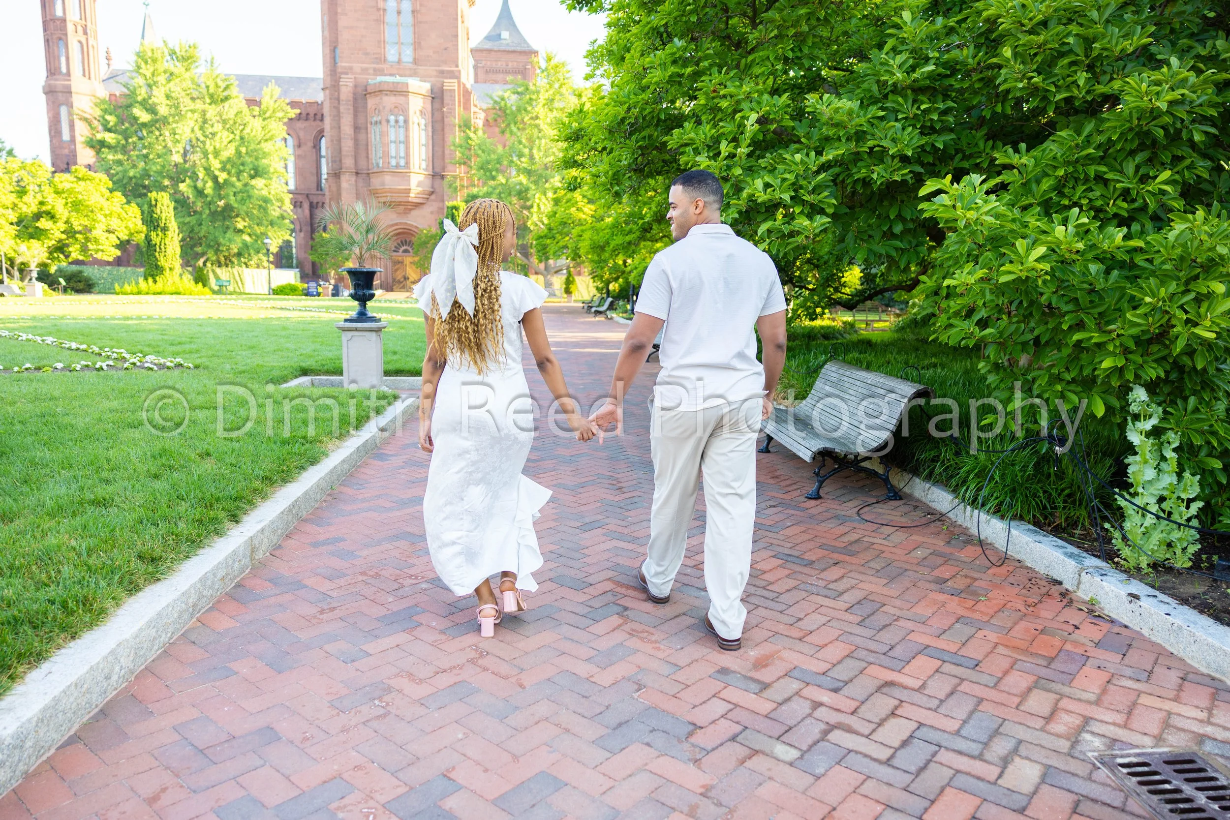 A couple holding hands and walking on a brick path in a park with green trees, benches, and a large brick church or cathedral building in the background during the daytime.