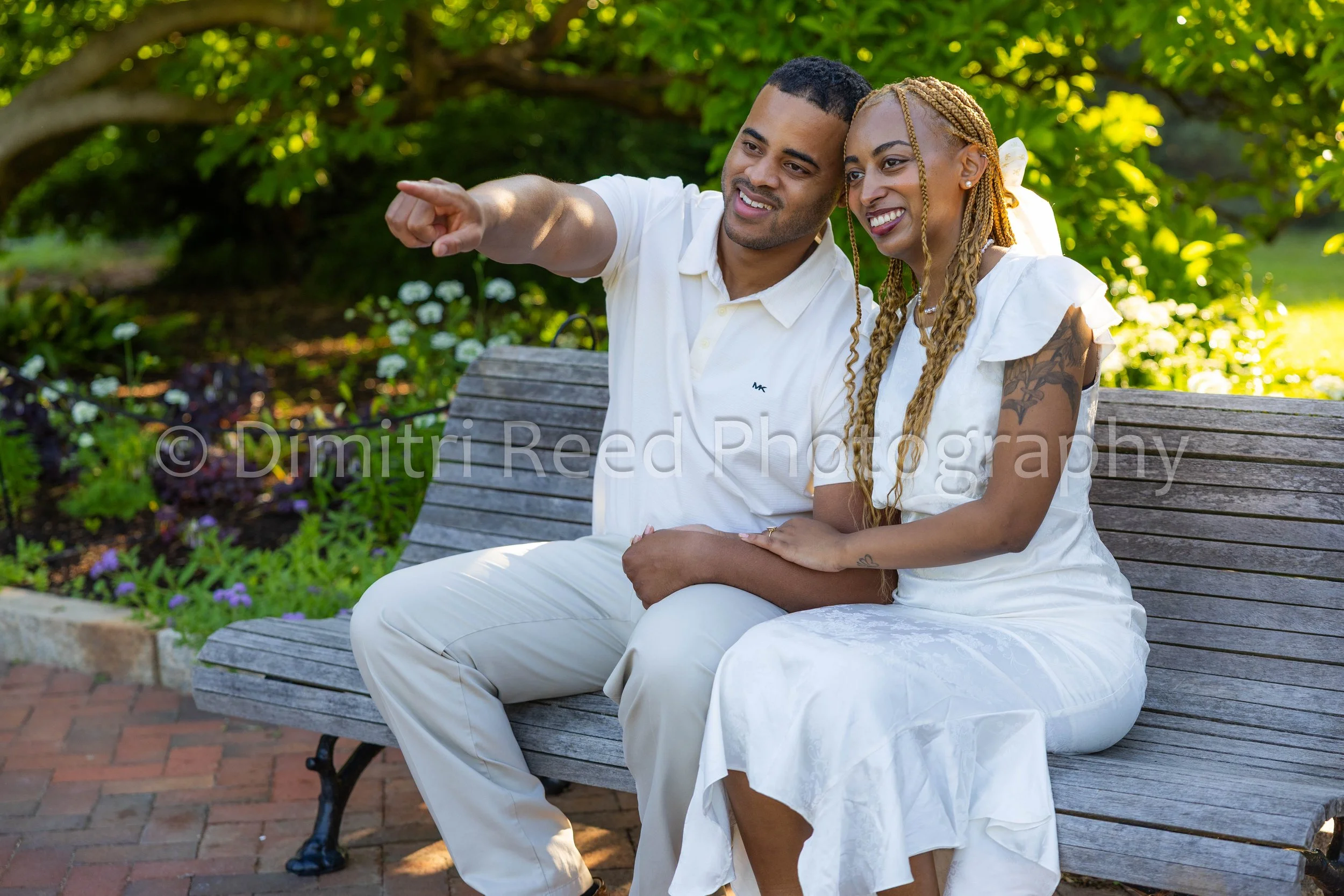 A smiling couple sitting on a park bench, outdoors with green trees in the background, pointing at something in the distance.