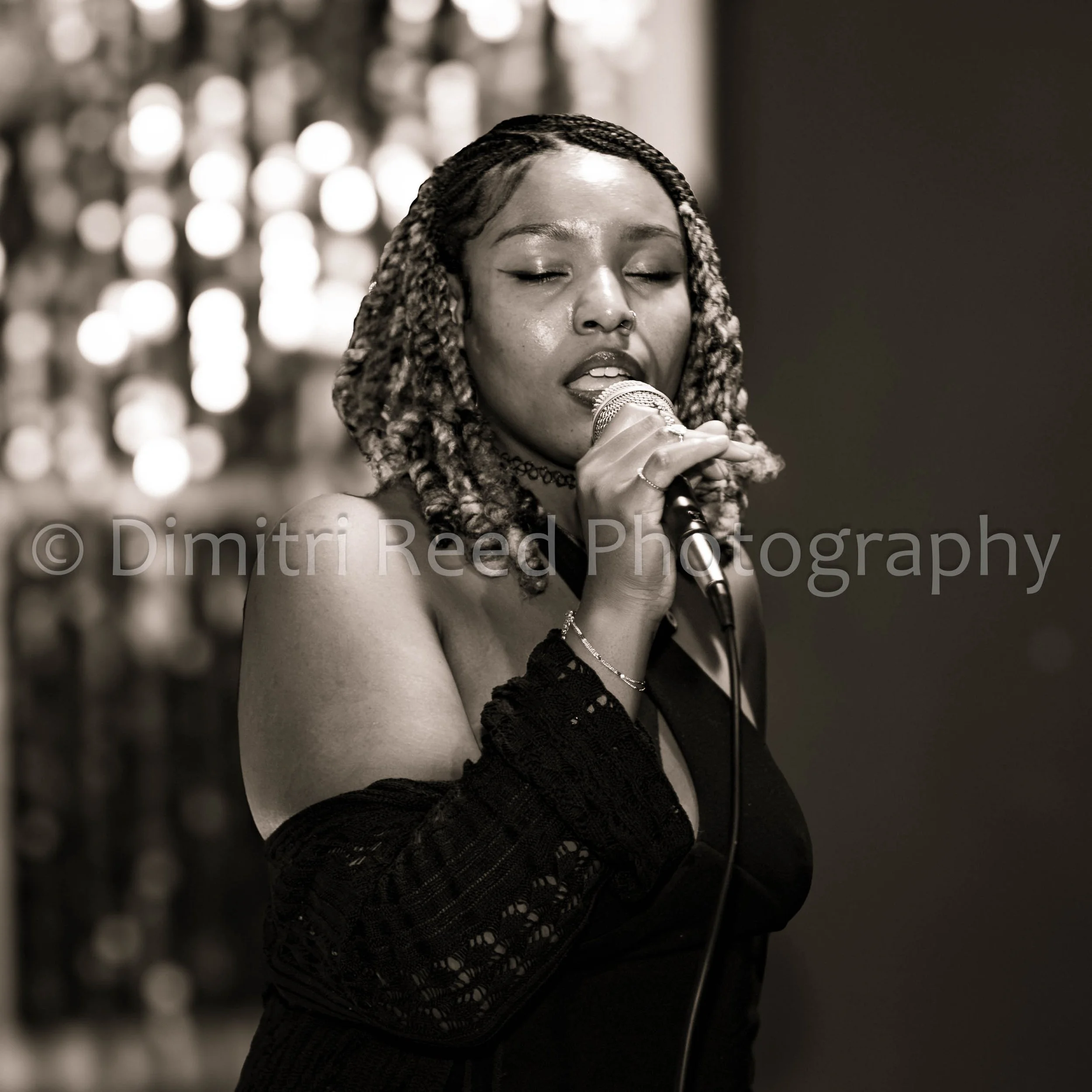 A woman with braided hair and a nose ring singing into a microphone with her eyes closed, wearing a black lace top and choker necklace, against a blurred background with circular lights.
