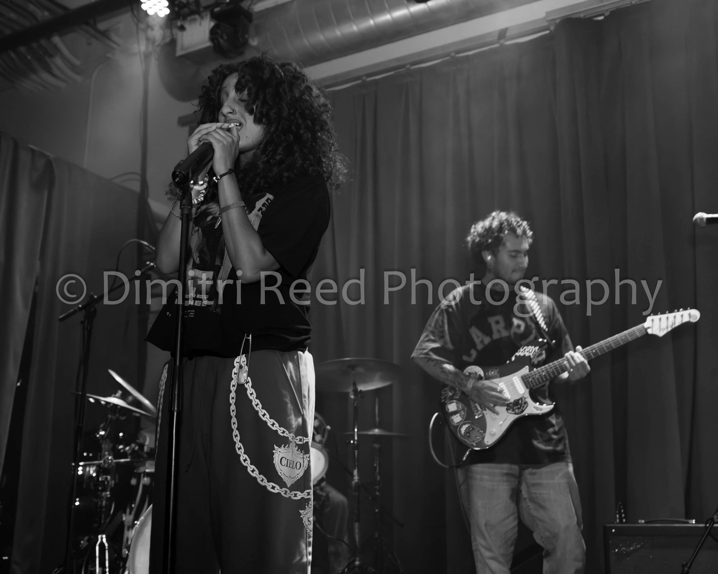 A woman singing into a microphone and a man playing an electric guitar on stage in a black and white photo.