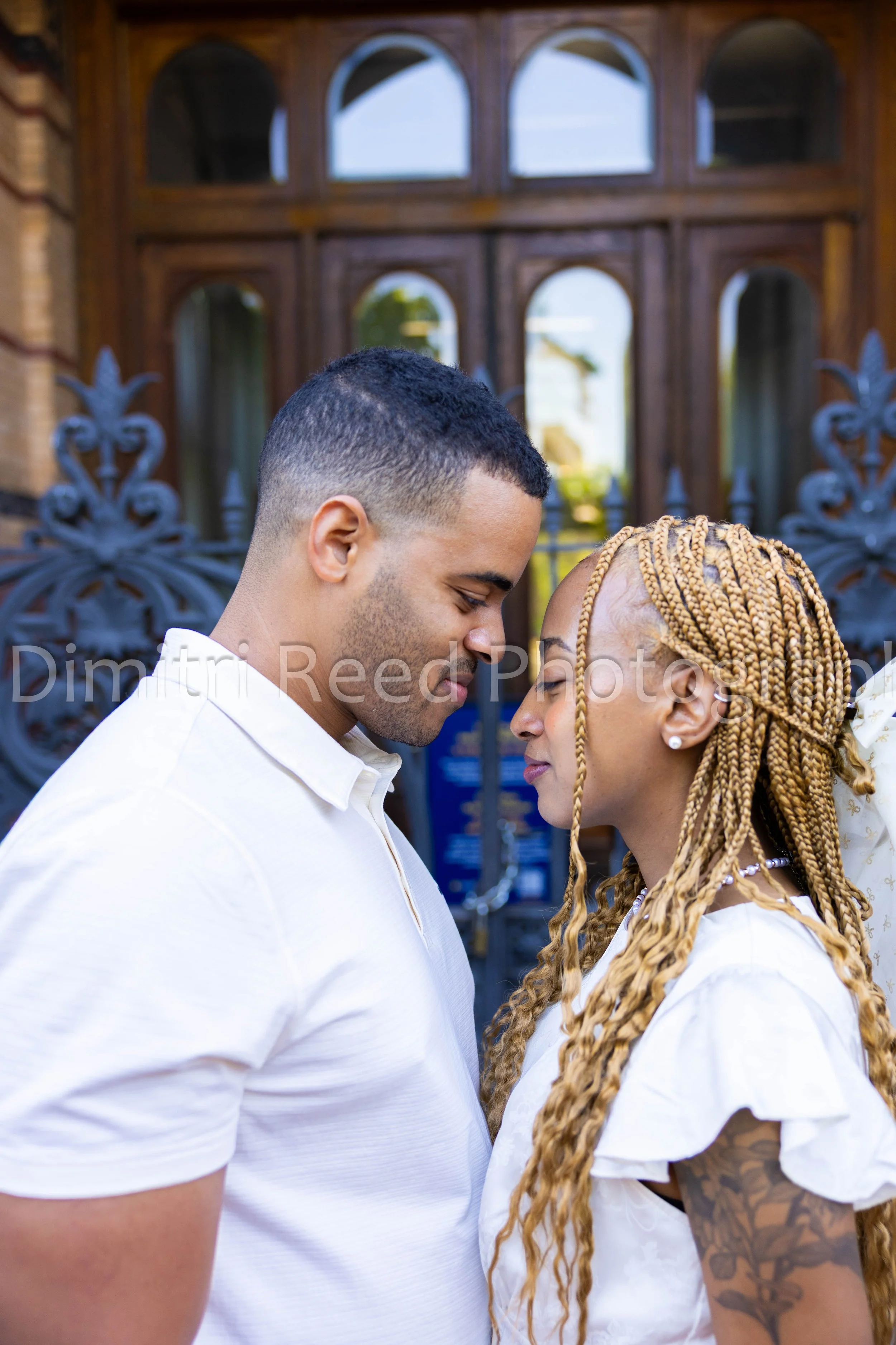 A young couple with their foreheads touching, eyes closed, standing closely outdoors in front of ornate wooden and metal gates.