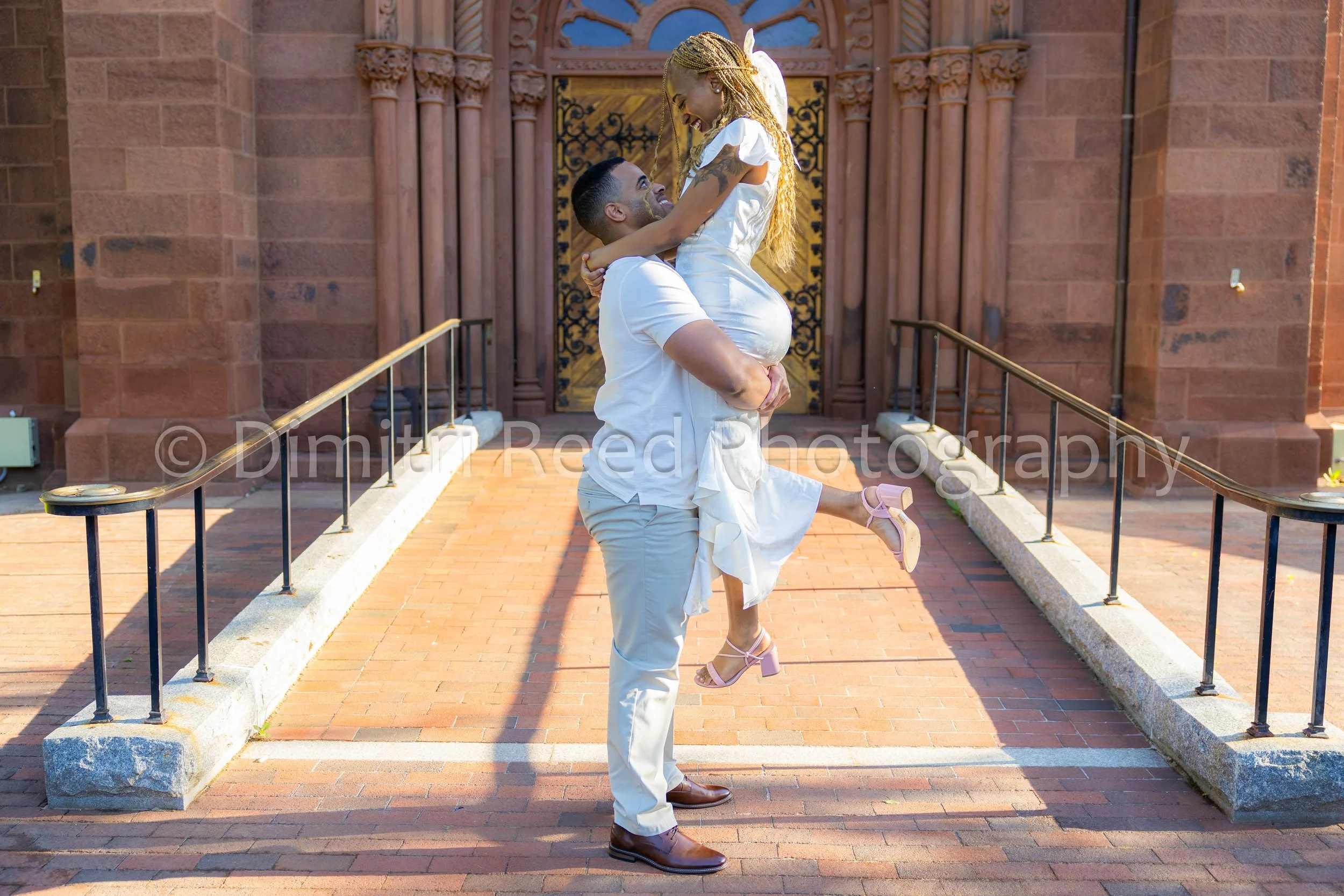 A man is lifting a woman in a white dress in front of a historic building with Gothic architecture.
