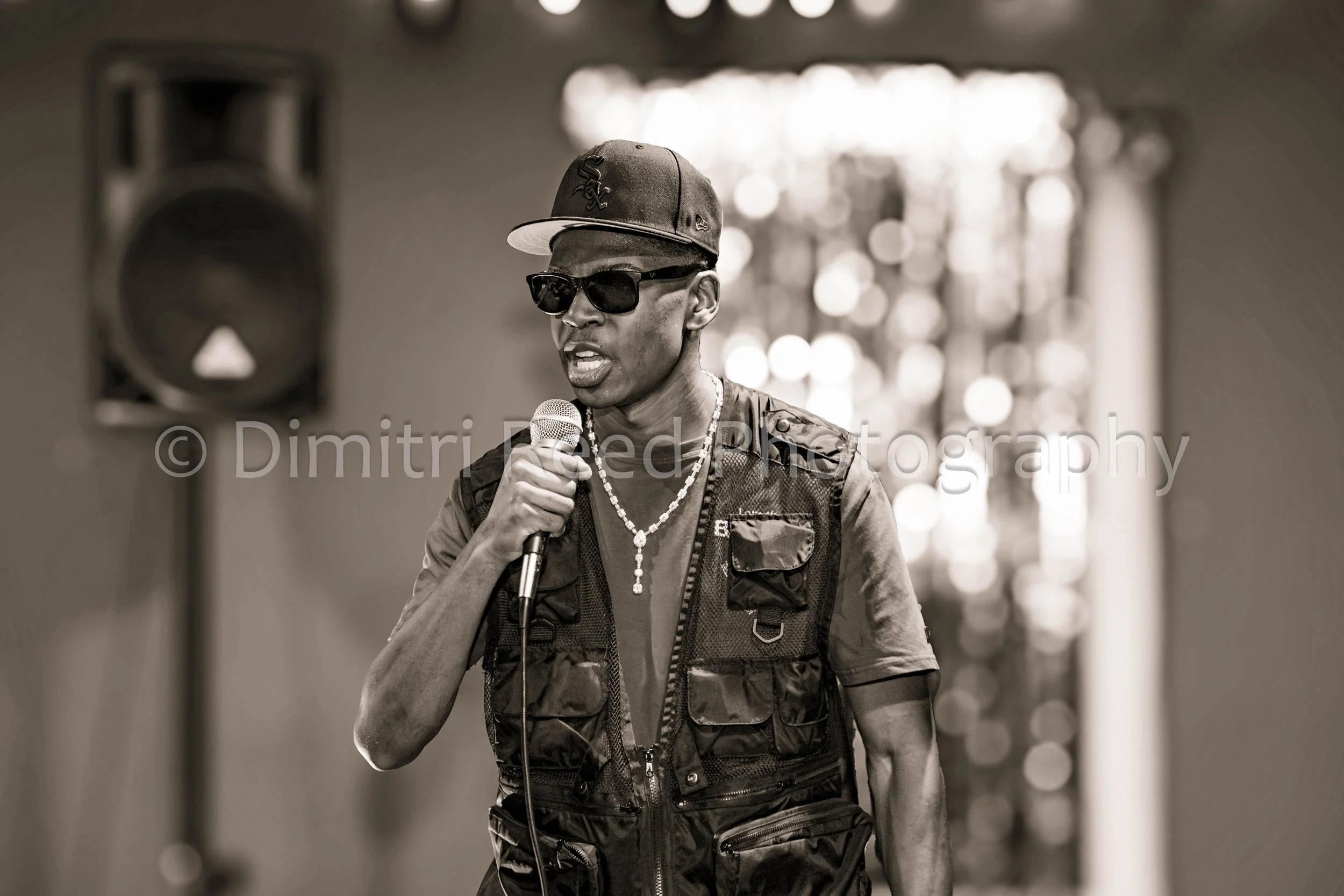 A man wearing sunglasses, a cap with the Chicago White Sox logo, a tactical vest, and jewelry, holding a microphone and speaking. The background has a blurred pattern of bright circular lights.