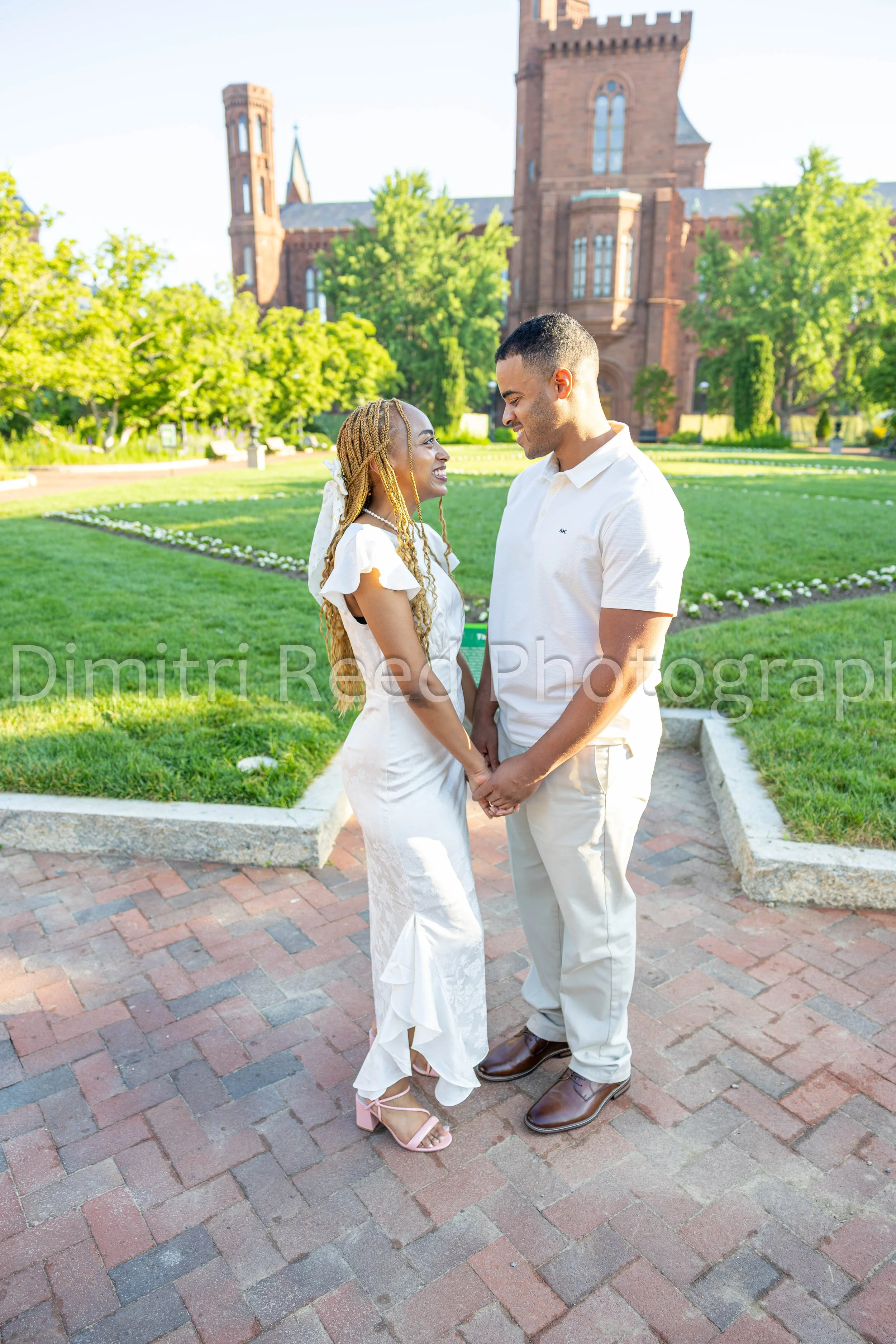A couple standing in a park, holding hands and looking lovingly into each other's eyes. The woman is wearing a white dress with ruffled sleeves and pink high-heeled sandals, and has long braided hair. The man is dressed in a white polo shirt, beige p