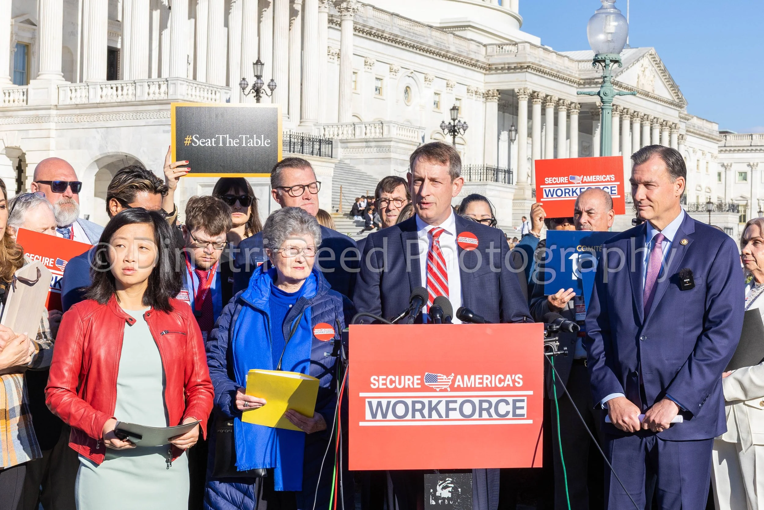 Group of people gathered outside the U.S. Capitol building during a rally or protest, with a man speaking at a podium that reads 'Secure America's Workforce' and signs that say '#SeatTheTable' and 'Secure America’s Workforce'.