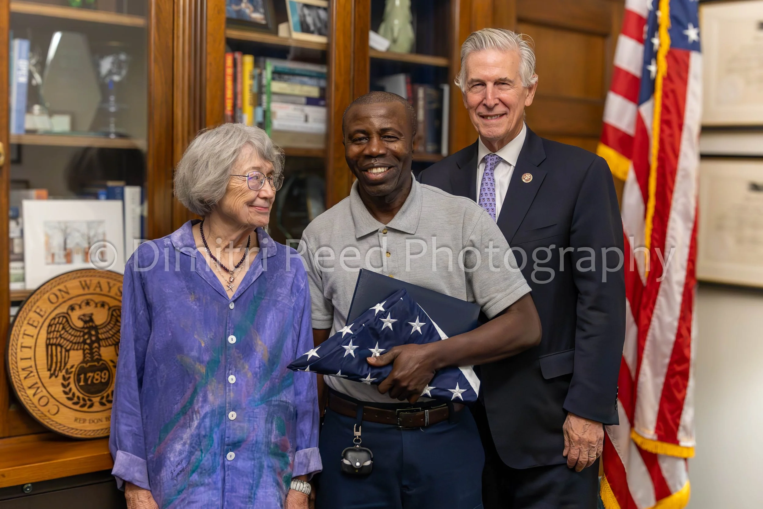 A man holding a folded American flag is smiling and standing between an elderly woman with gray hair and glasses and a man with white hair in a suit. They are indoors with a bookshelf and American flags in the background.
