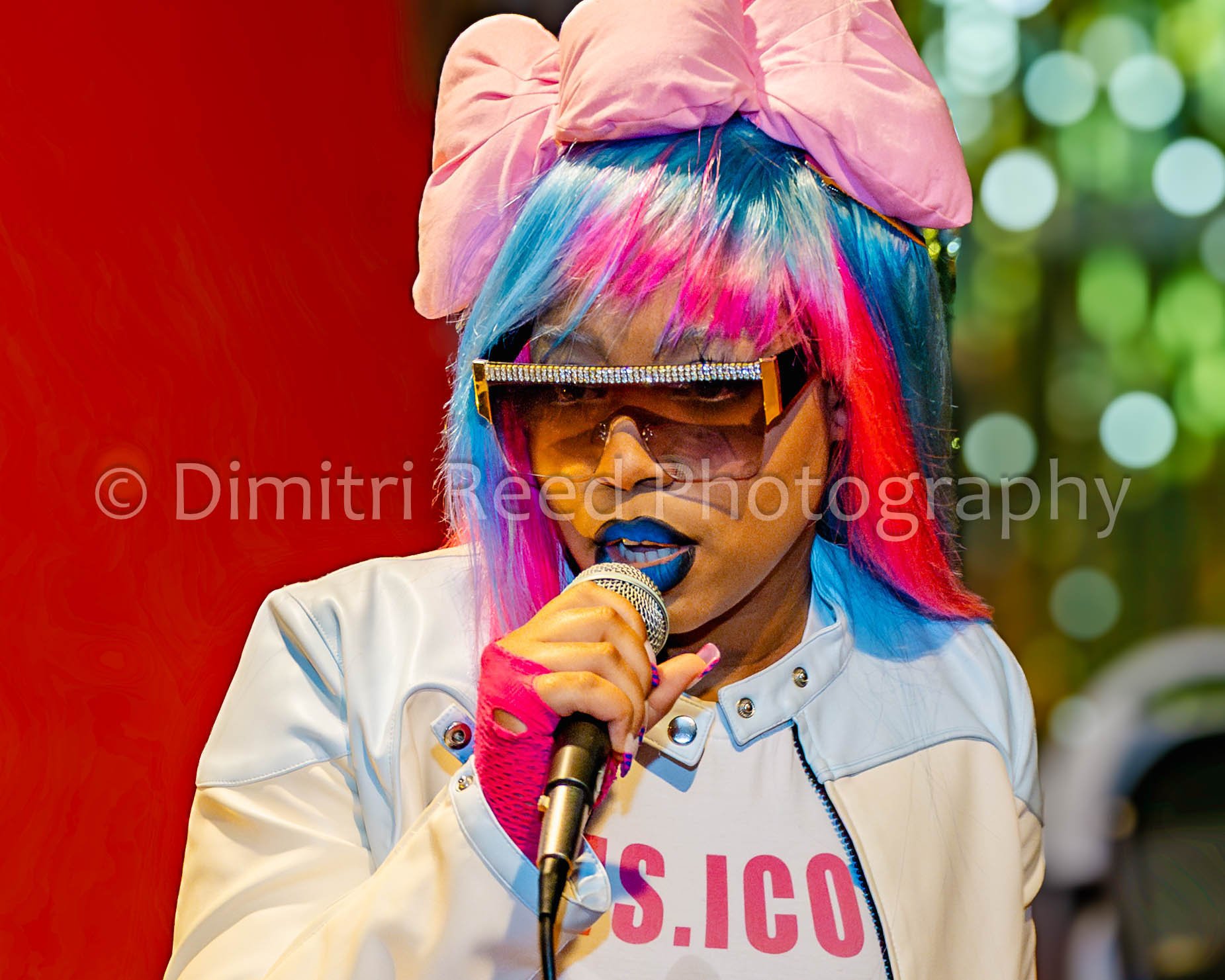 A person with rainbow-colored hair, wearing a pink bow headwrap, sunglasses with rhinestones, a white leather jacket, and pink gloves, holding a microphone and speaking.
