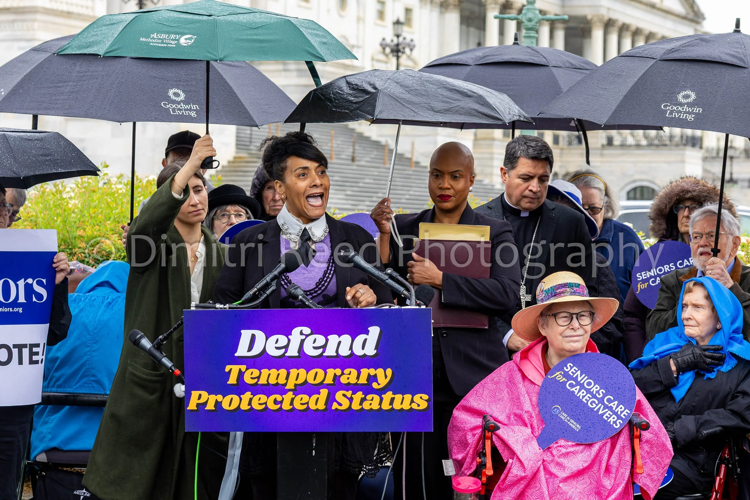 A group of people gathered outdoors holding umbrellas during a protest or rally, with speakers addressing the crowd from a podium. The main speaker is a woman speaking into a microphone, with various signs and banners visible, including a blue sign w