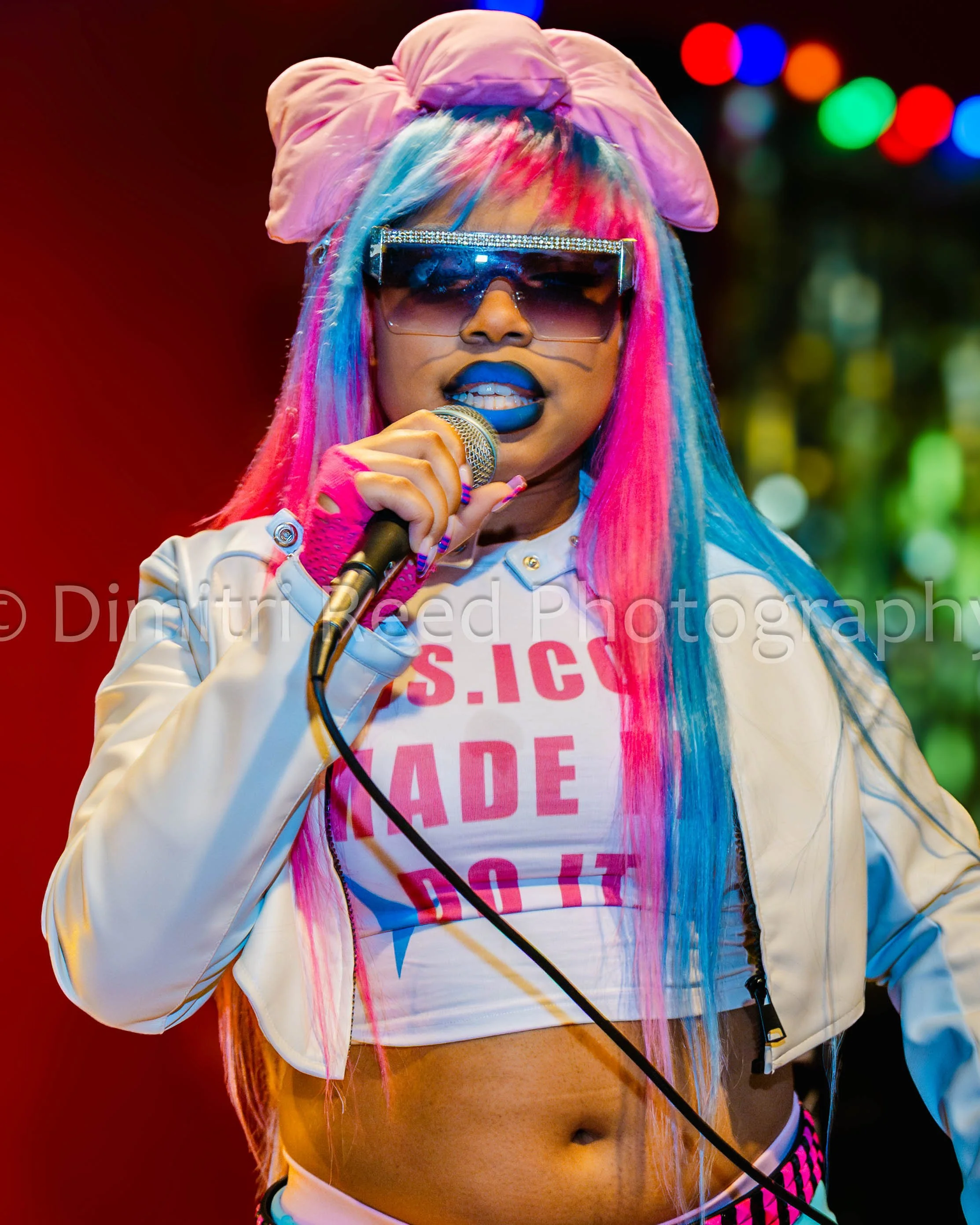 A woman with colorful dyed hair, wearing large sunglasses with decorative rhinestones, a pink bow headband, and a white shirt with pink text, singing into a microphone against a background of vibrant, colorful lights.