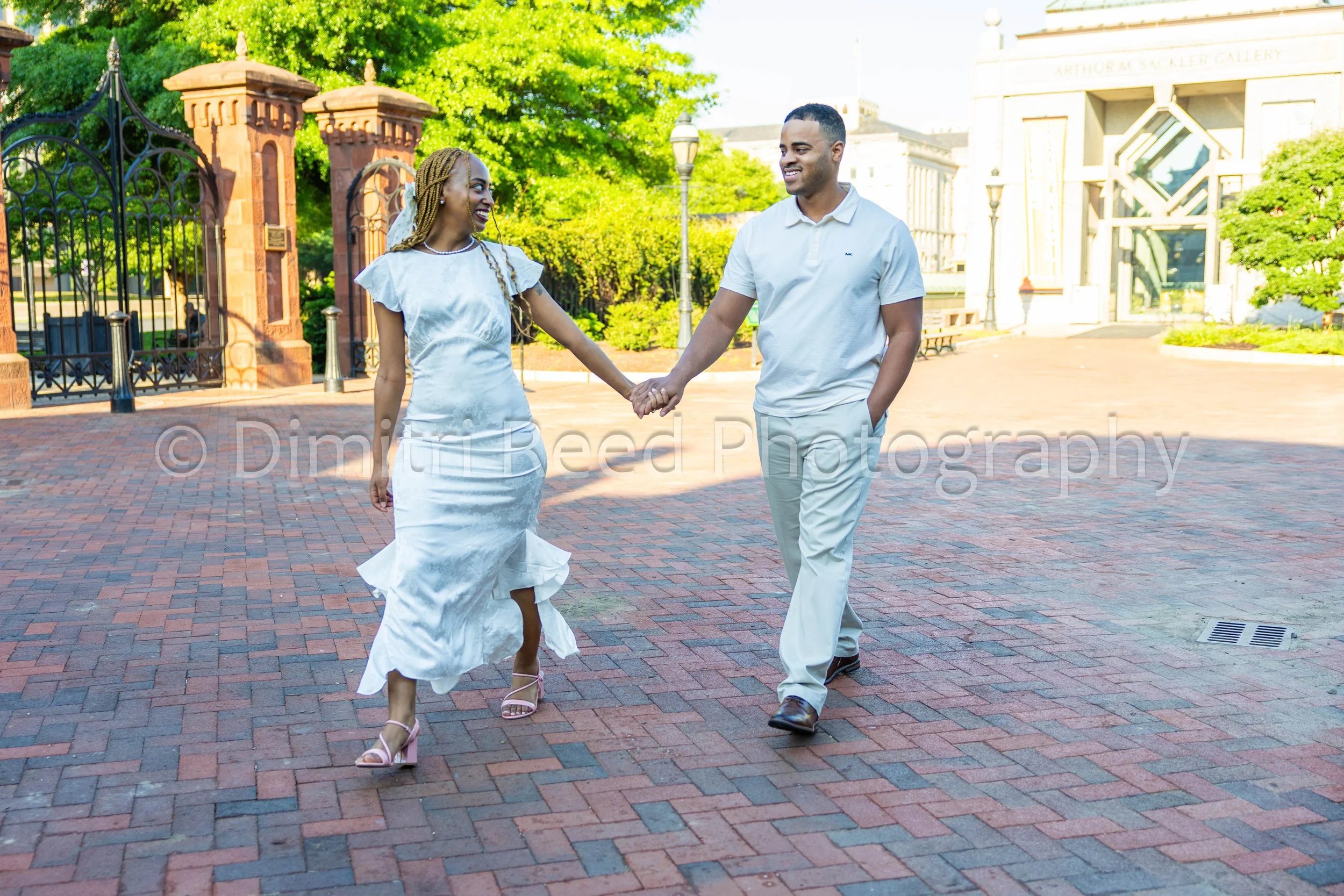 A couple holding hands and dancing outside in front of a historic building on a sunny day.