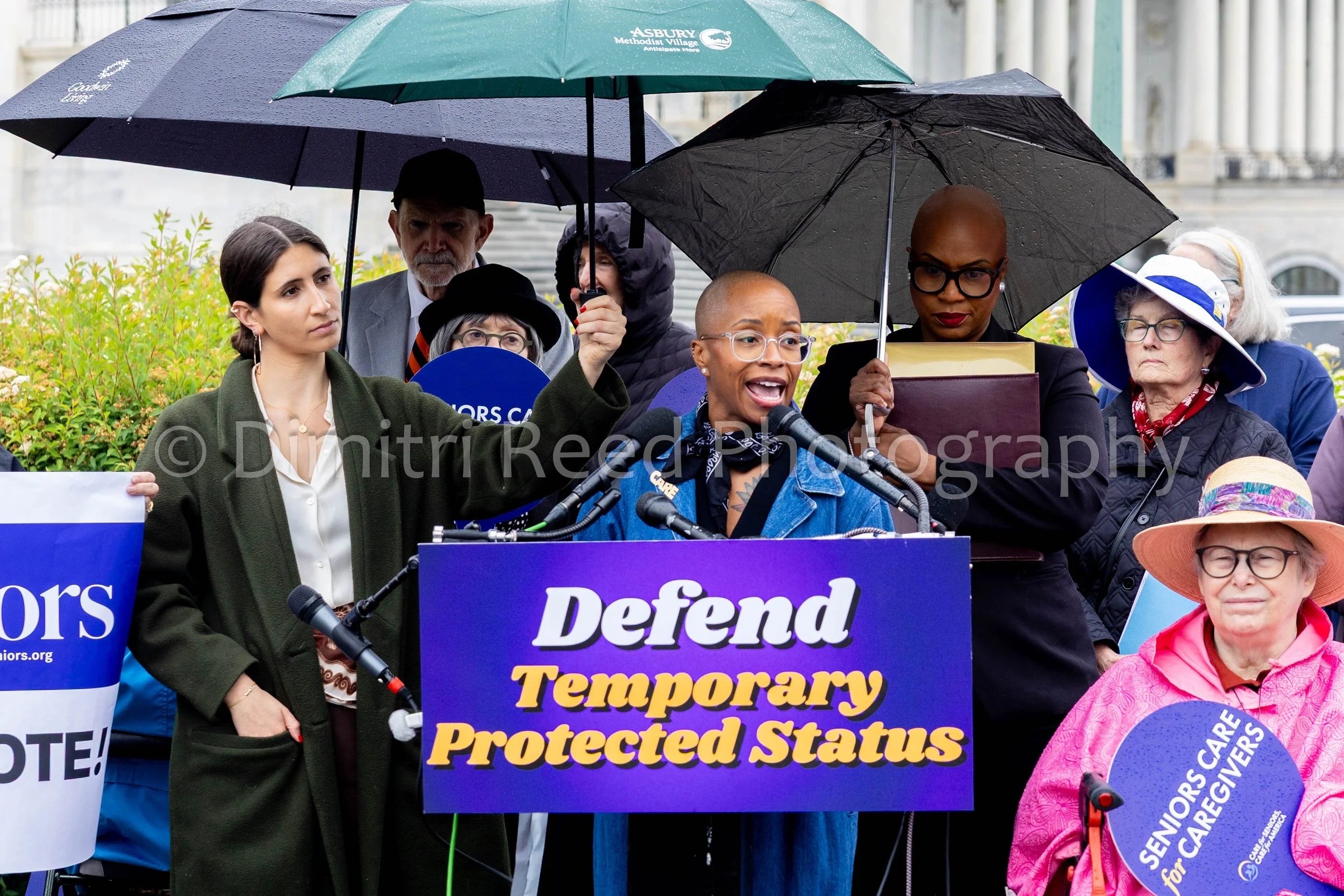 A diverse group of people holding umbrellas and standing behind a podium at a protest or rally. The sign on the podium reads, 'Defend Temporary Protected Status.' Some individuals are wearing jackets and hats, and there are banners and signs related 