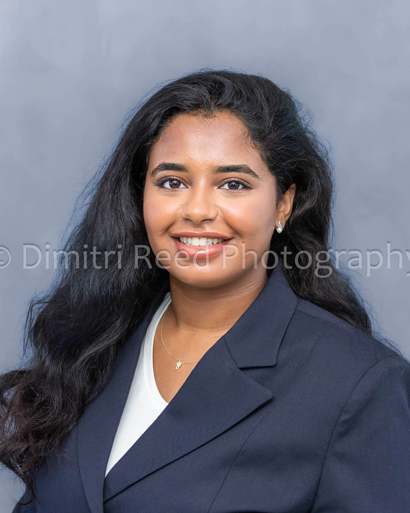 Headshot of a woman with long, wavy black hair, wearing pearl earrings, a navy blazer, and a white top, smiling against a neutral gray background.