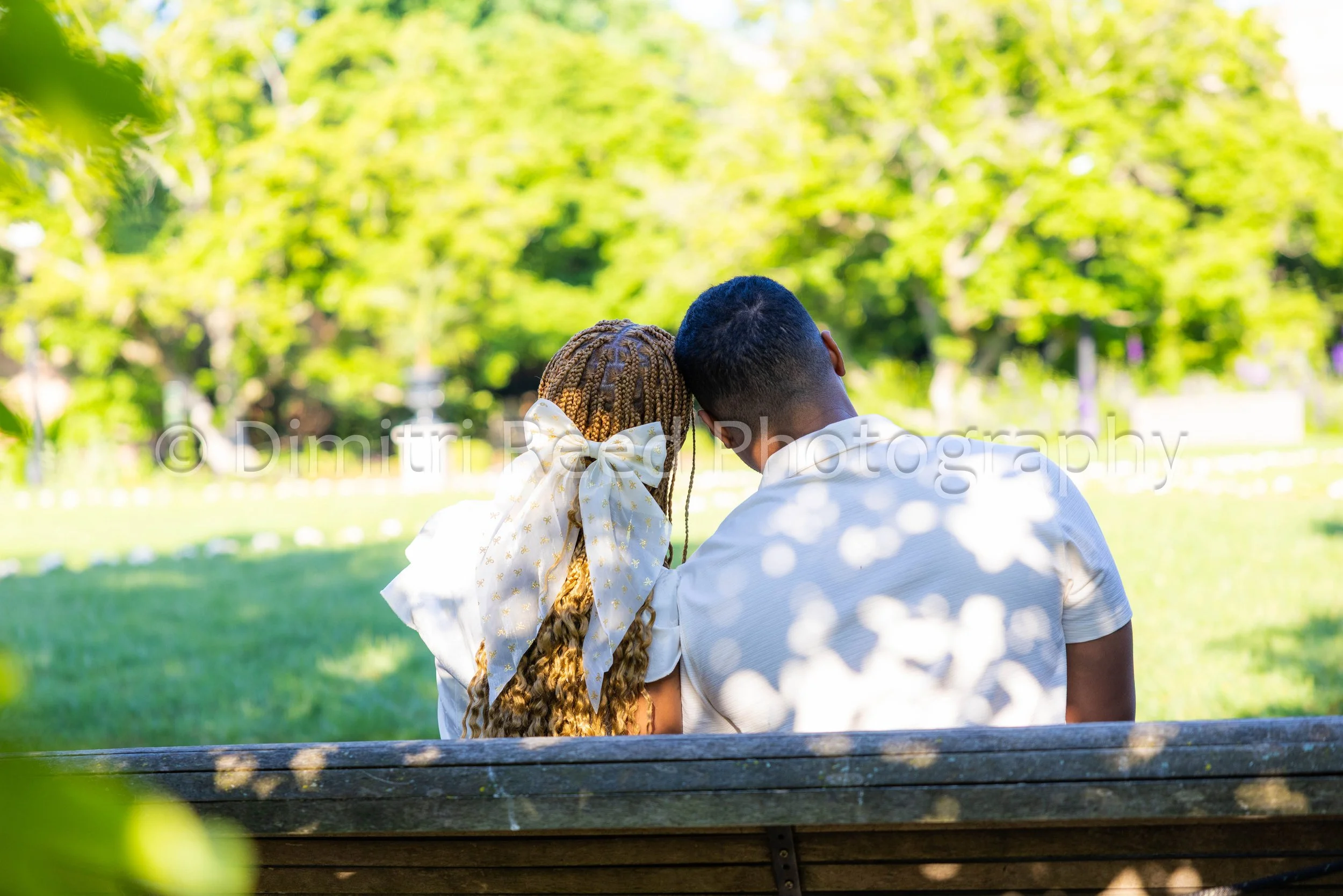 A couple sitting on a park bench under trees, with their heads close together, enjoying a sunny day outside.