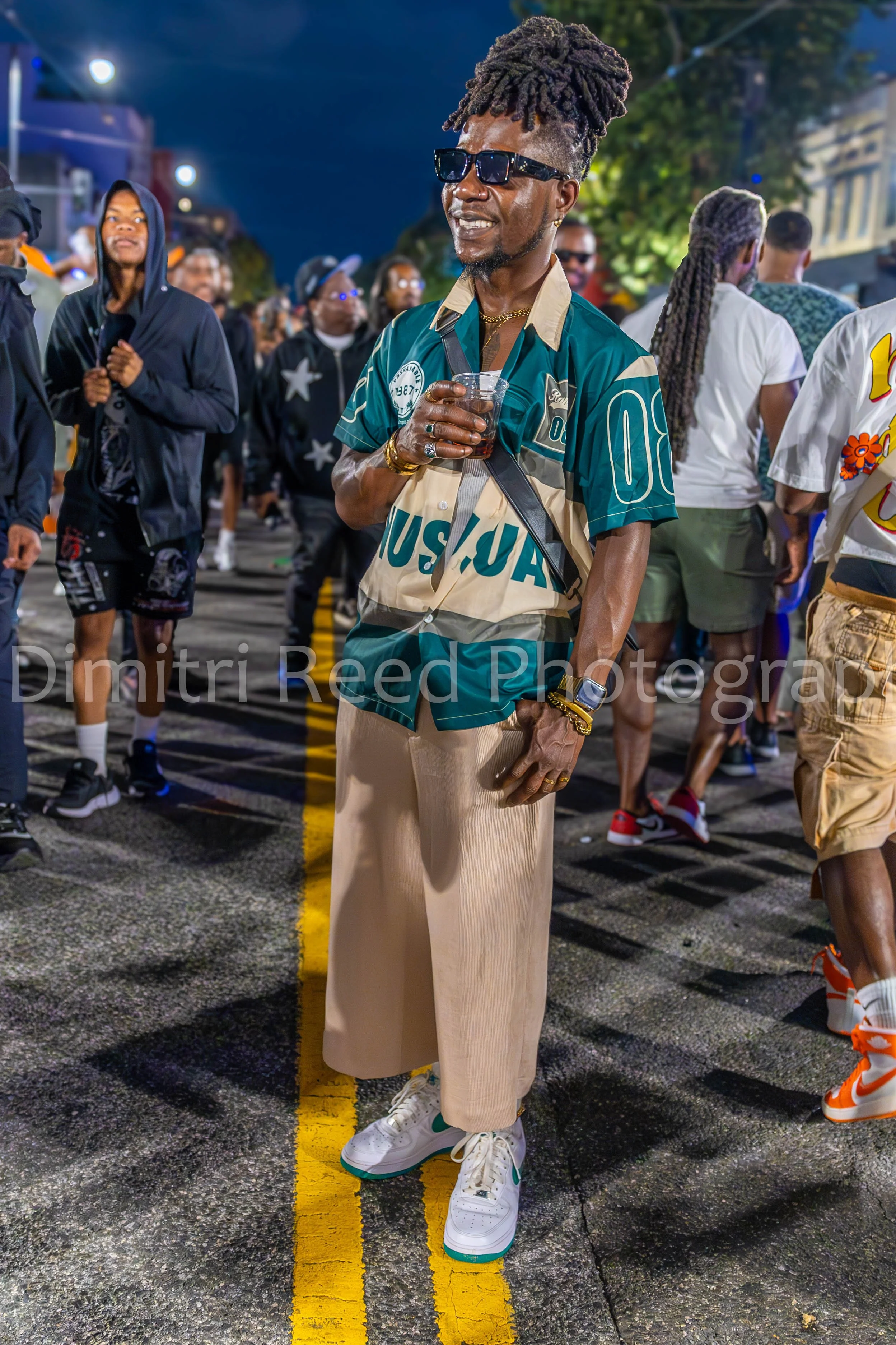 A man in a vintage U.S. Army uniform shirt and beige pants, holding a drink in one hand, standing on a city street at night surrounded by a crowd. He wears sunglasses, a gold watch, gold bracelets, and has a tall hairstyle with locks. People in casua