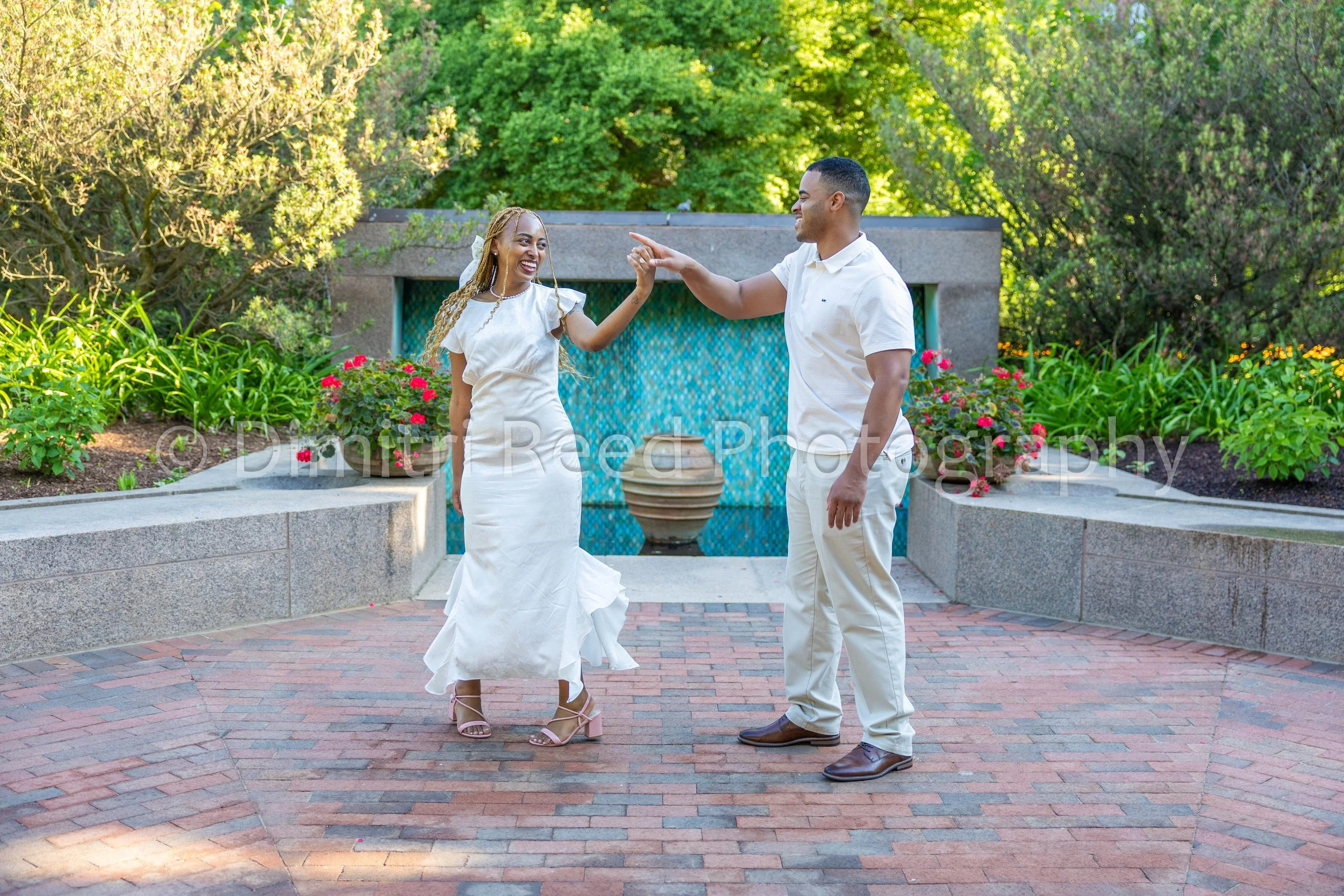 A woman in a white dress and heels dancing with a man in a white shirt and beige pants outdoors in a garden with green trees and pink flowers.