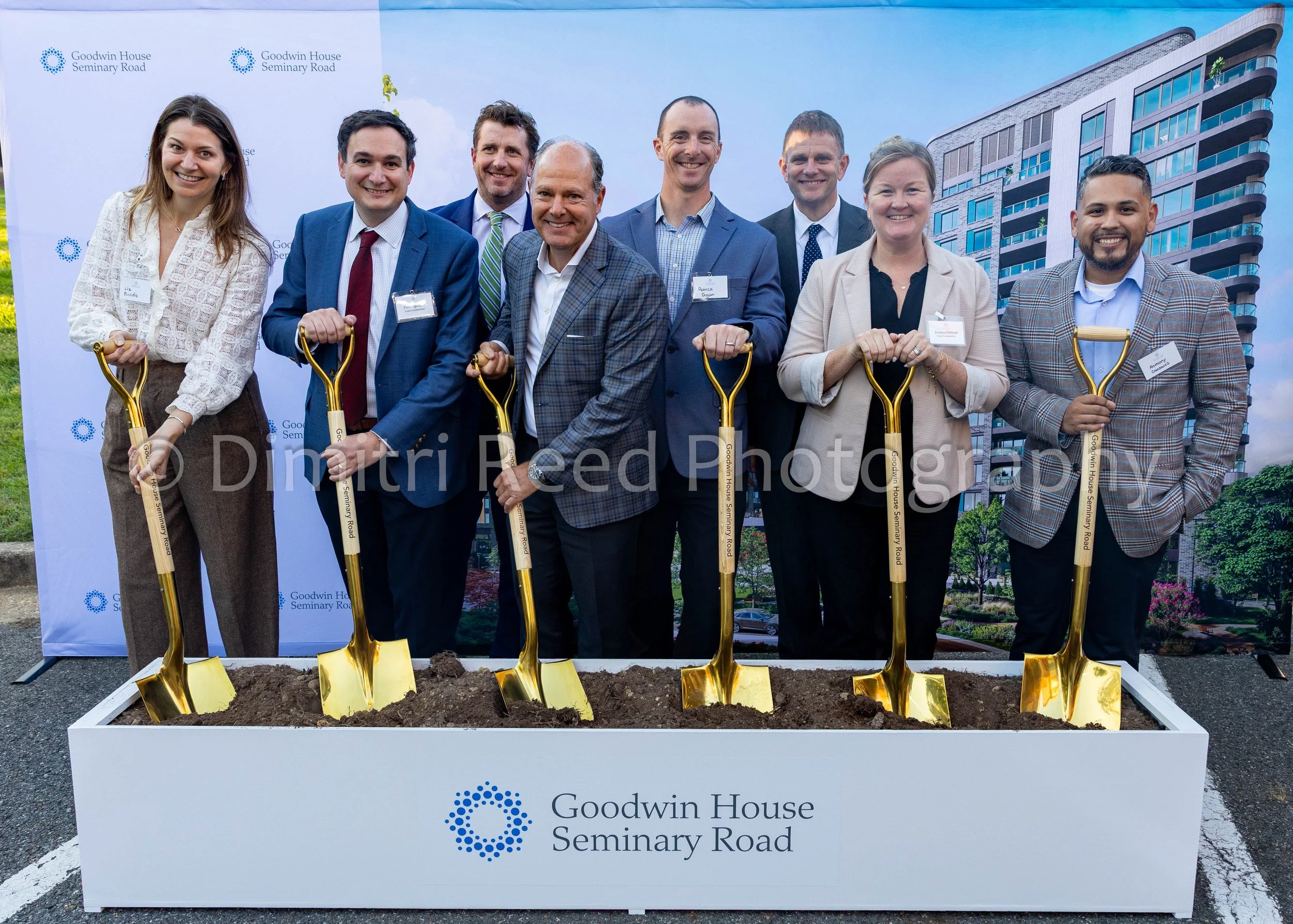 Group of nine people, seven men and two women, smiling and holding small gold shovels, standing in front of a sign that says 'Goodwin House Seminary Road,' with a newly planted tree in the background.