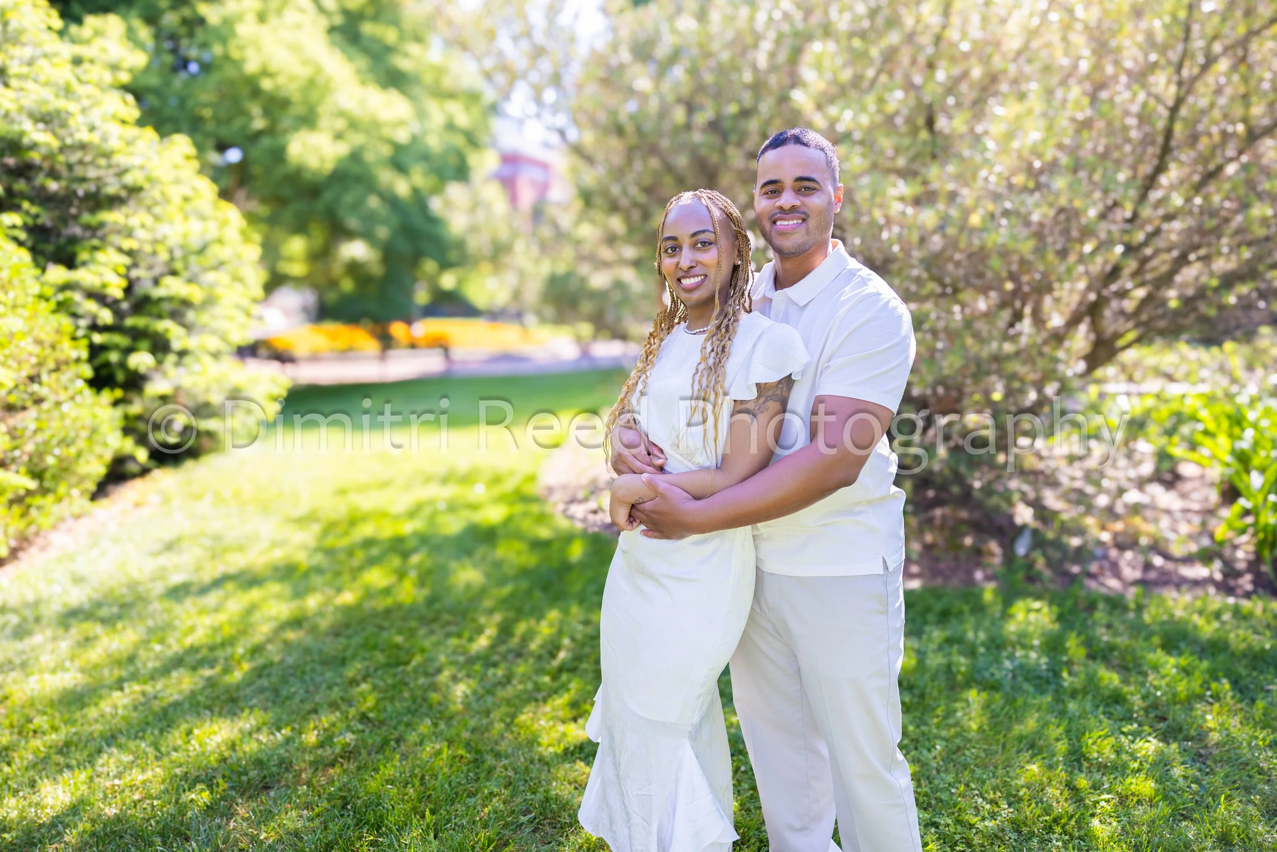 A happy couple embracing outdoors in a lush, green park with trees and sunlight filtering through the leaves.
