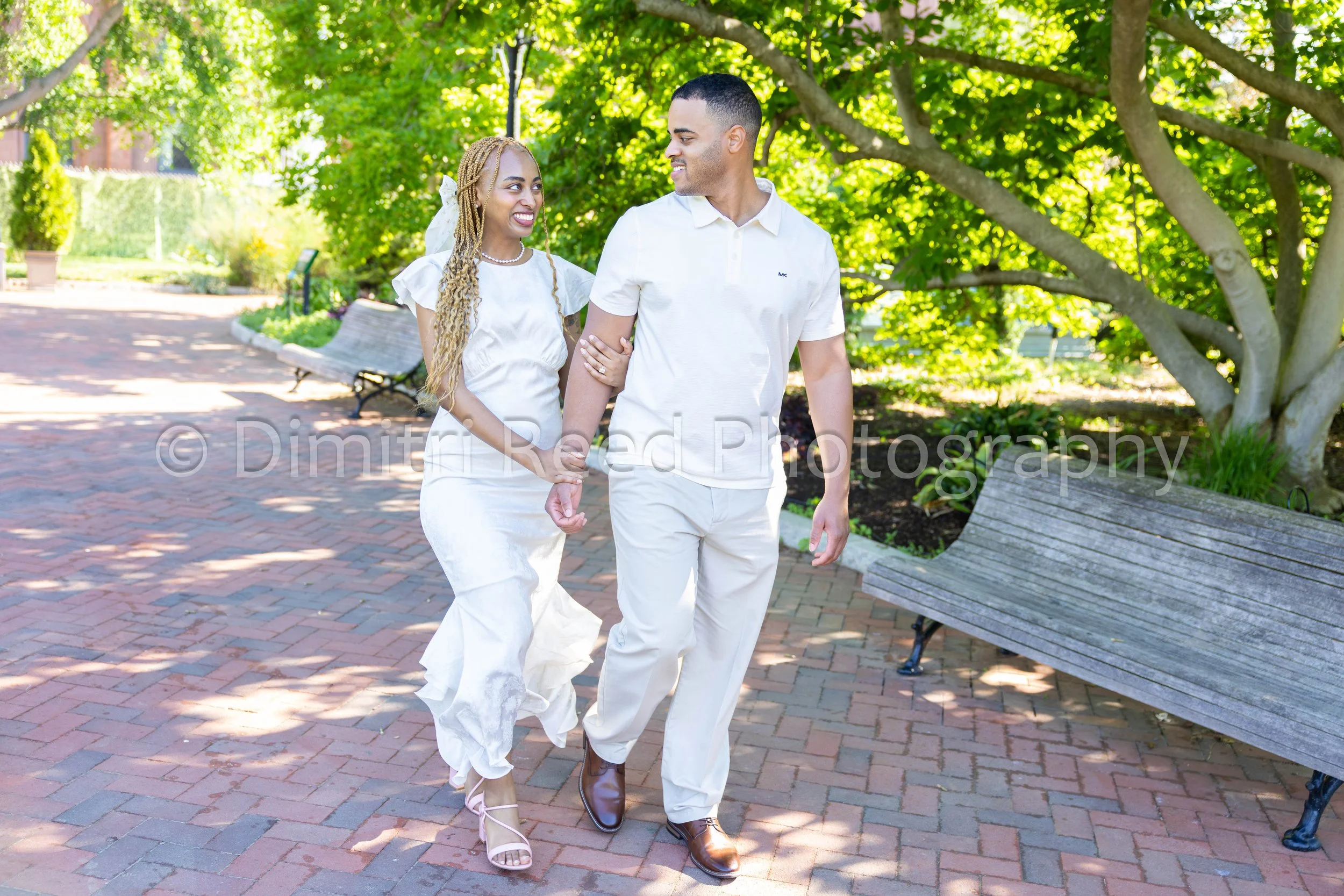 A smiling couple walking arm in arm in a park on a sunny day, surrounded by green trees and benches.
