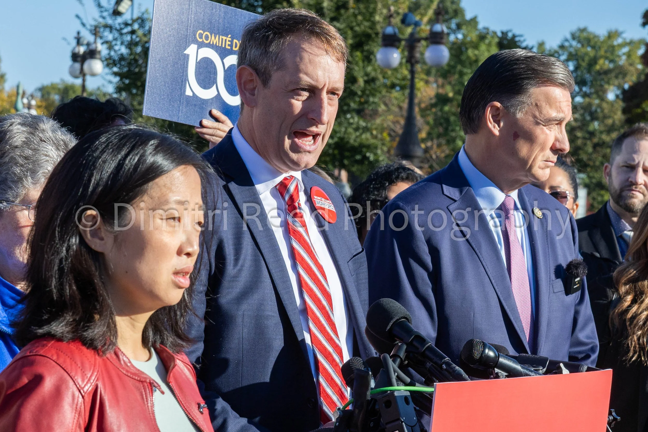 Group of people, including a woman with dark hair in a red jacket and men in suits, at an outdoor event with microphones and a banner in the background.