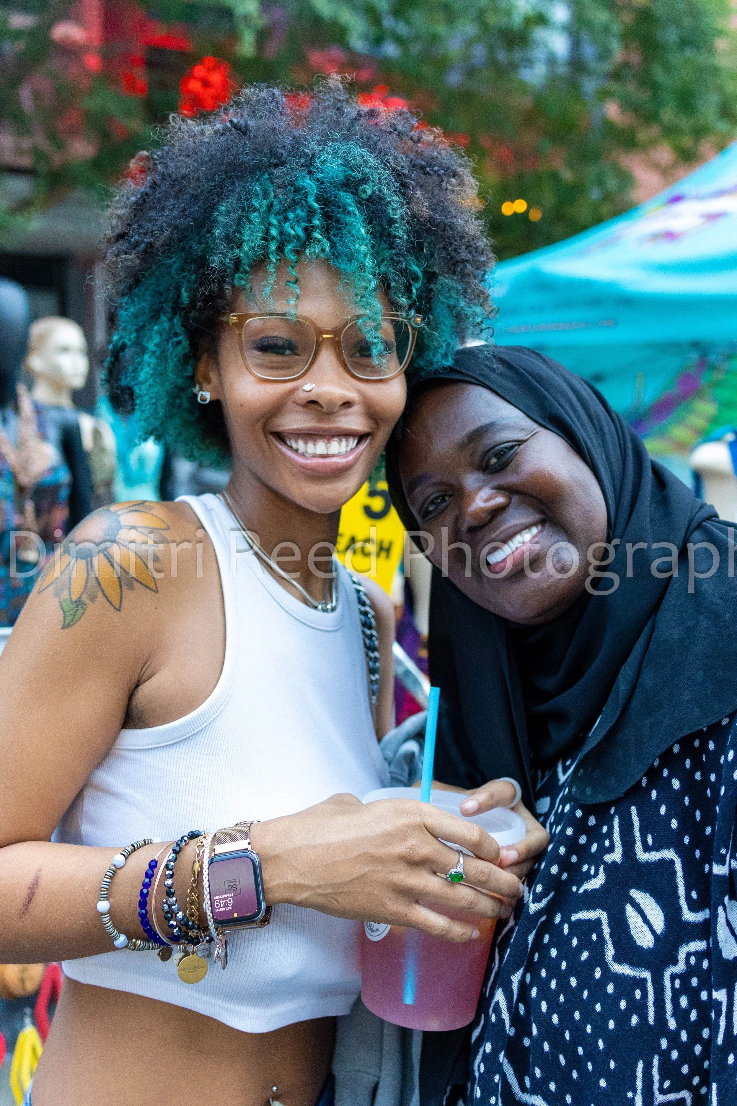 Two women smiling and hugging at an outdoor event, with tents and colorful decorations in the background.