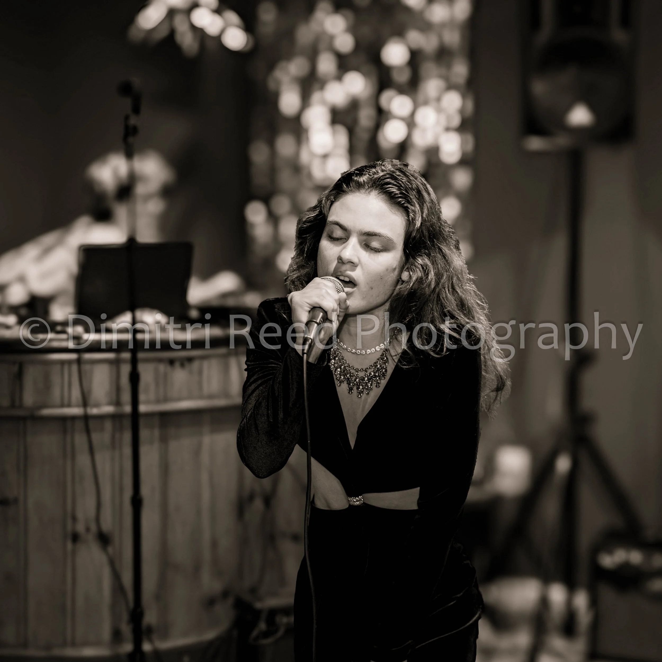 A woman with long curly hair singing into a microphone in a dimly lit setting, with blurred lights in the background.