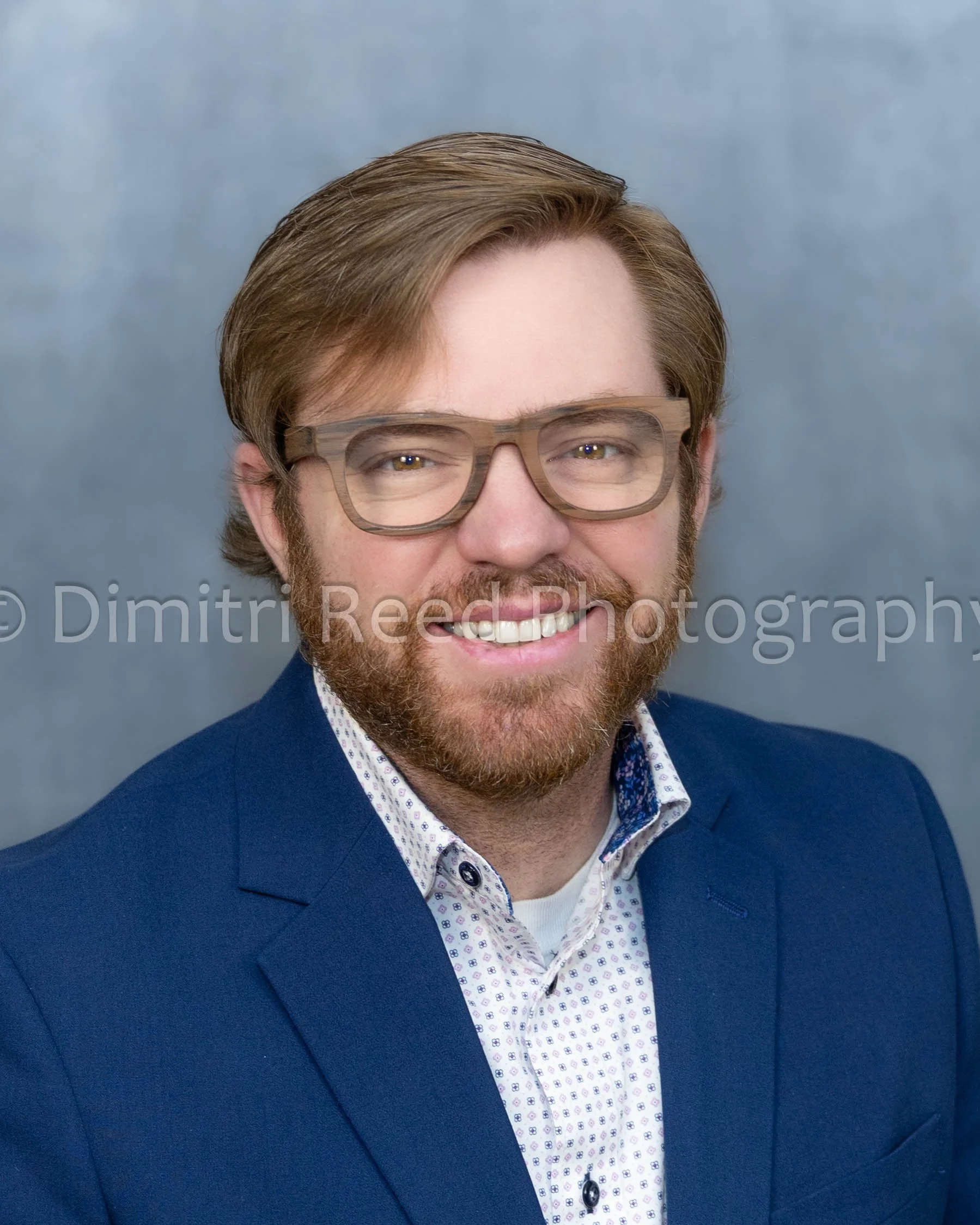 A man with brown hair and glasses is smiling, dressed in a navy blue suit with a patterned white shirt, against a plain background.