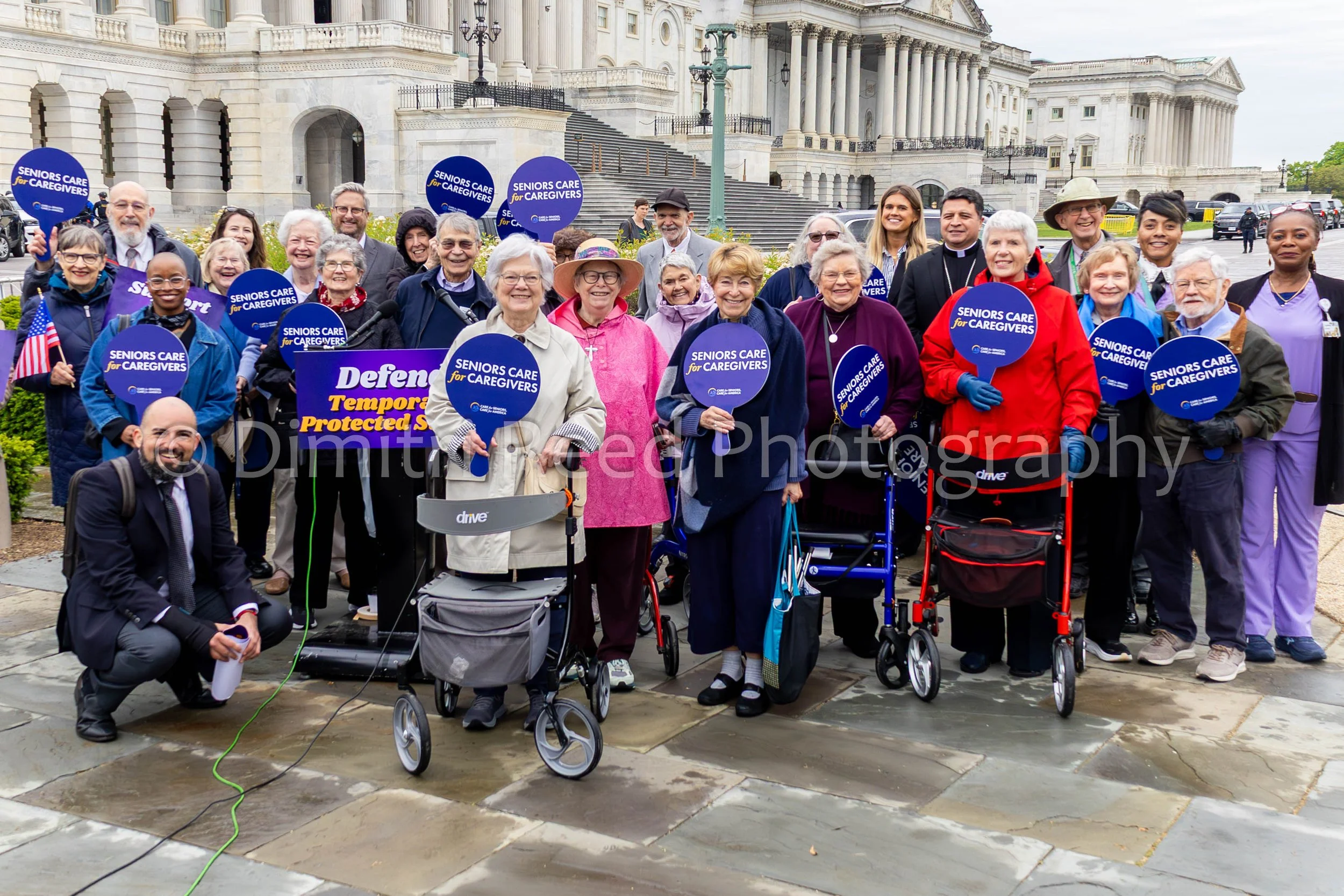 Group of diverse seniors and caregivers holding signs that read "Seniors Care for Caregivers" in front of a government building, with steps and columns, during a daytime rally.