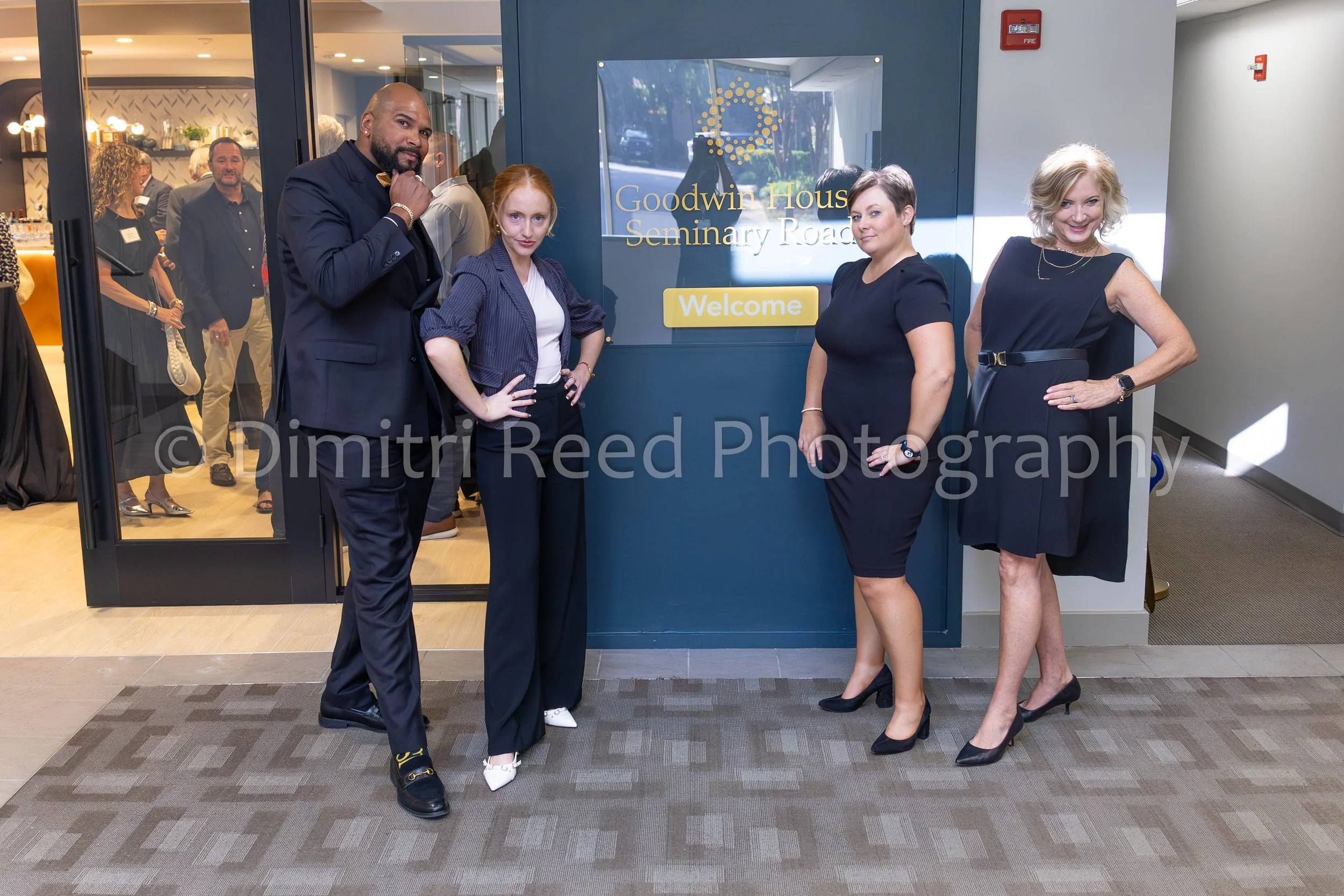 Four women and one man standing in front of a sign that reads "Goodwin House Seminary Road" with a yellow "Welcome" button. The women are dressed in formal or business attire, and the man is wearing a suit and bow tie. Behind them, other people are s